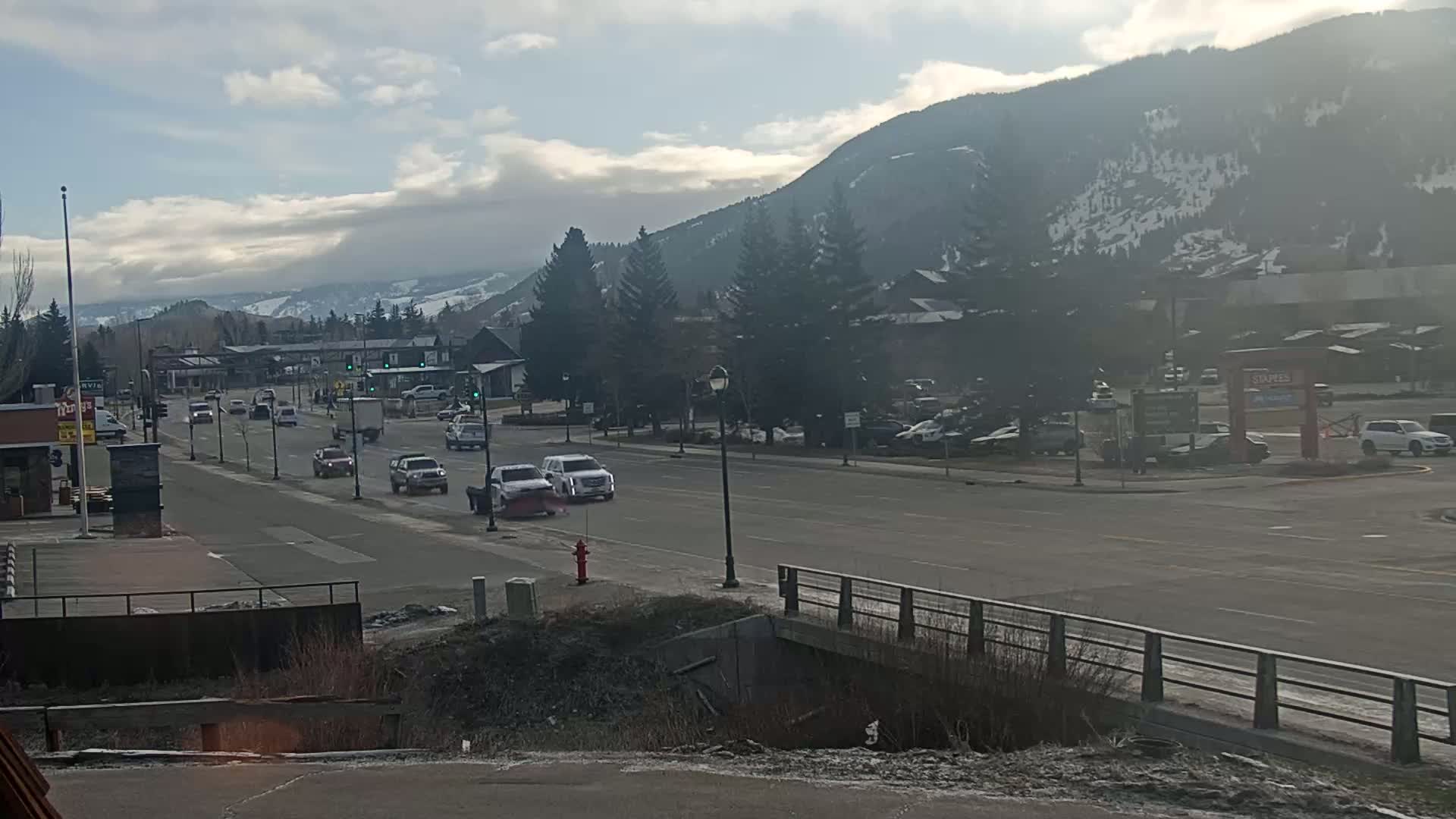 A partly cloudy day shows a lightly trafficked road leading to a snow-capped mountain range with buildings and trees lining the road.