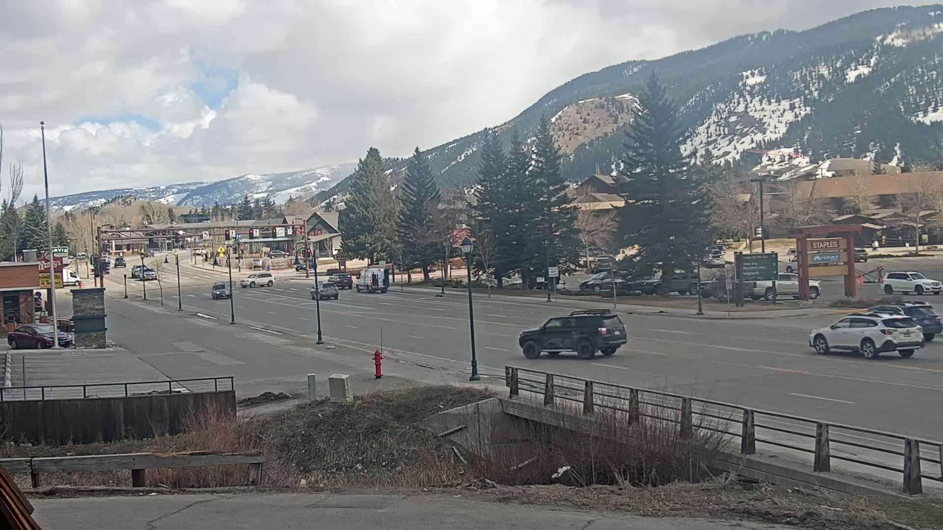 A partly cloudy day reveals a mountain town scene with a main road lined with vehicles, buildings, and evergreen trees, backed by snow-dusted mountains.