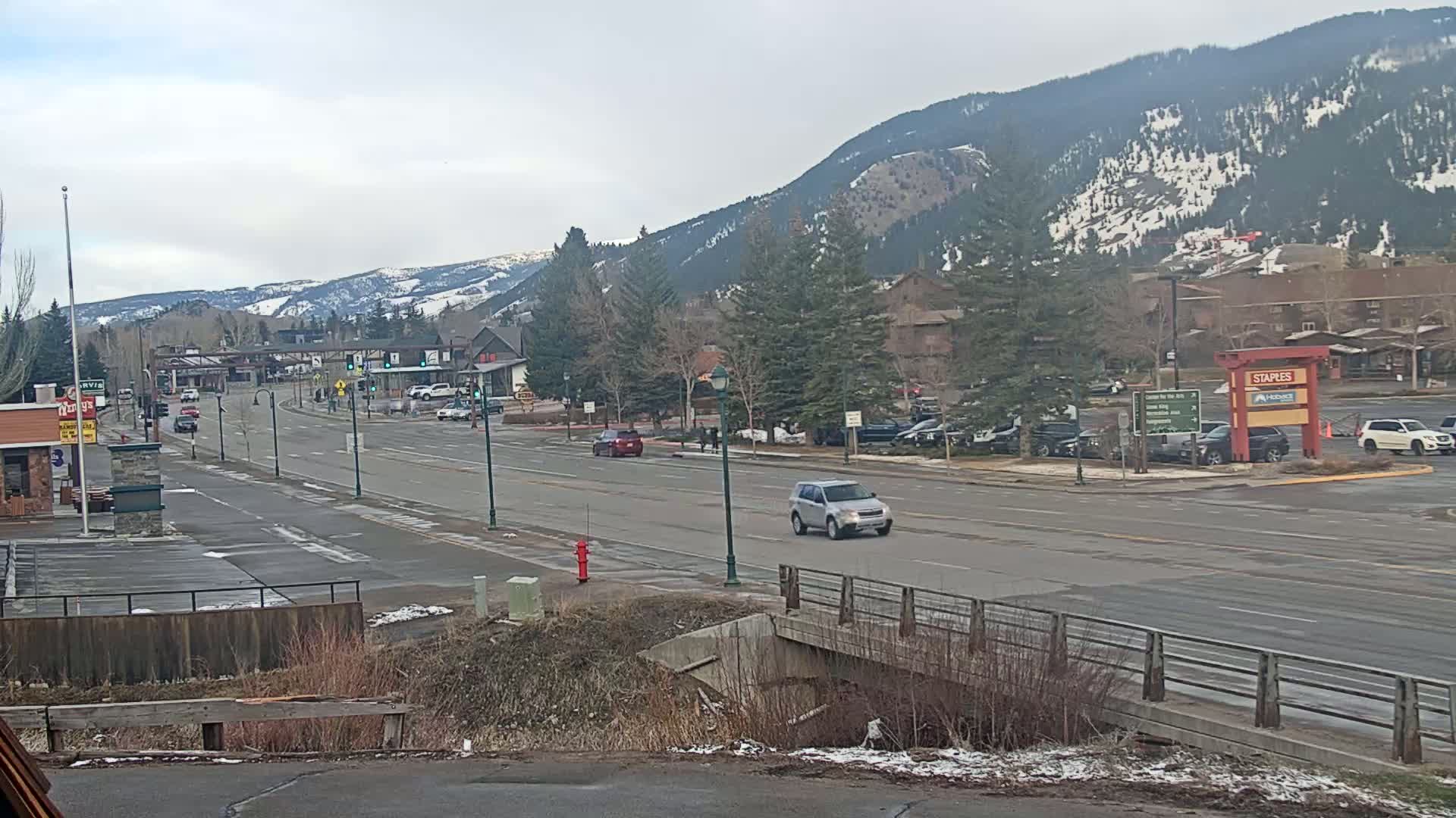 A mostly clear day reveals a mountain town scene with a multi-lane road, sparse traffic, and snow-dusted mountains in the background.