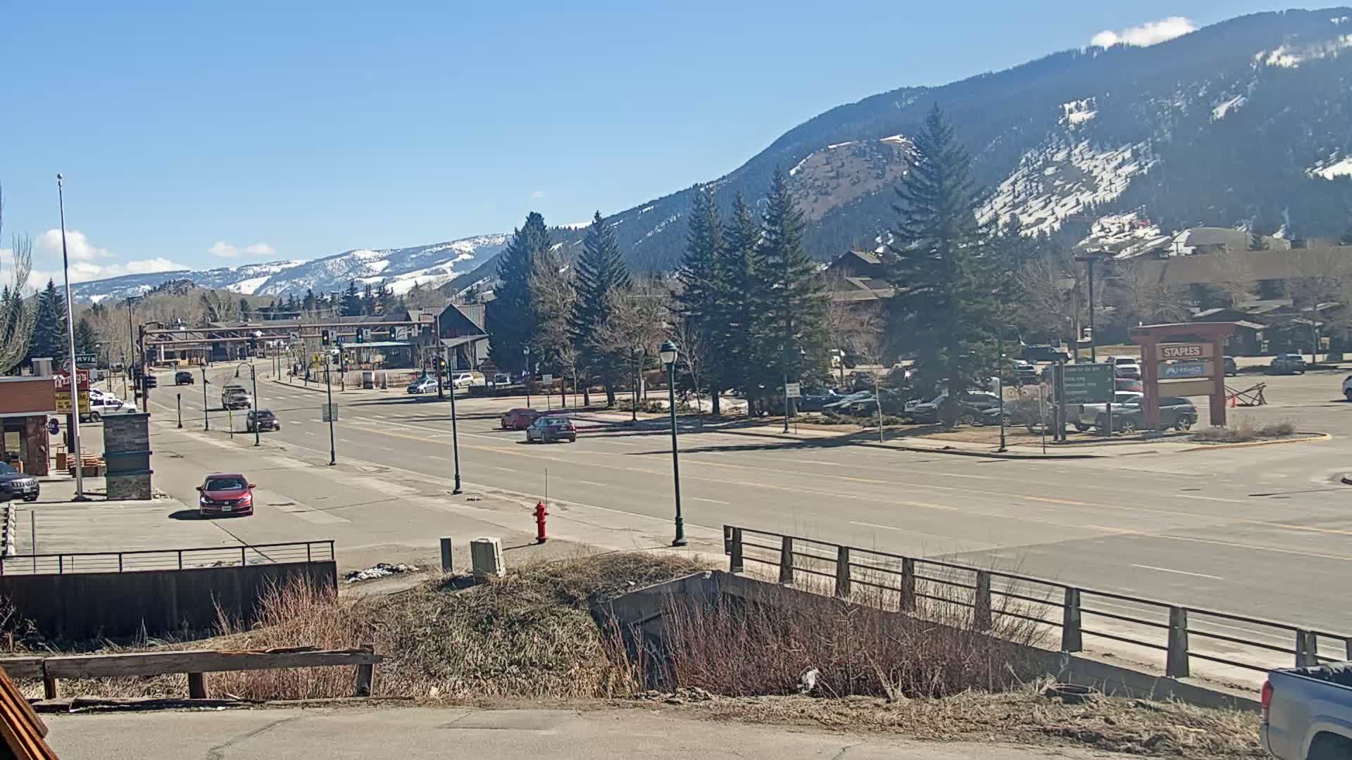 A sunny day reveals a town nestled in a valley, with snow-capped mountains in the background and sparse traffic on a wide road.