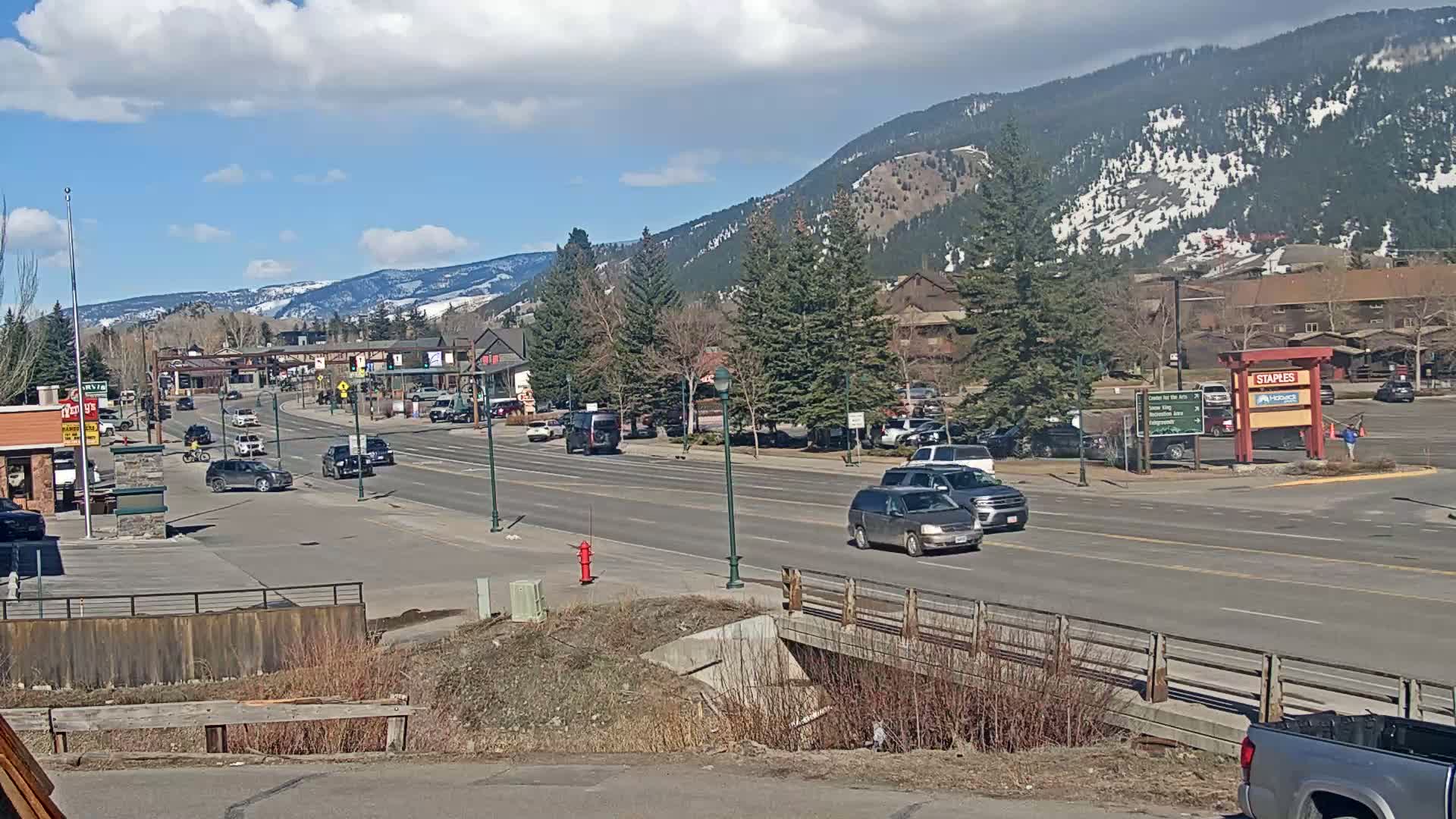 A sunny day reveals a mountain town scene with cars driving on a main road that passes by shops and buildings, backed by snow-capped mountains.