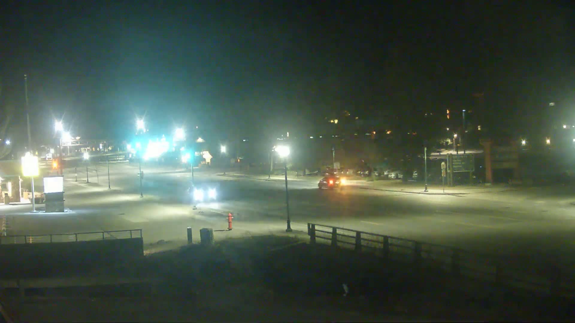 A nighttime view of a mostly empty road intersection with several cars and streetlights, illuminated under hazy conditions.