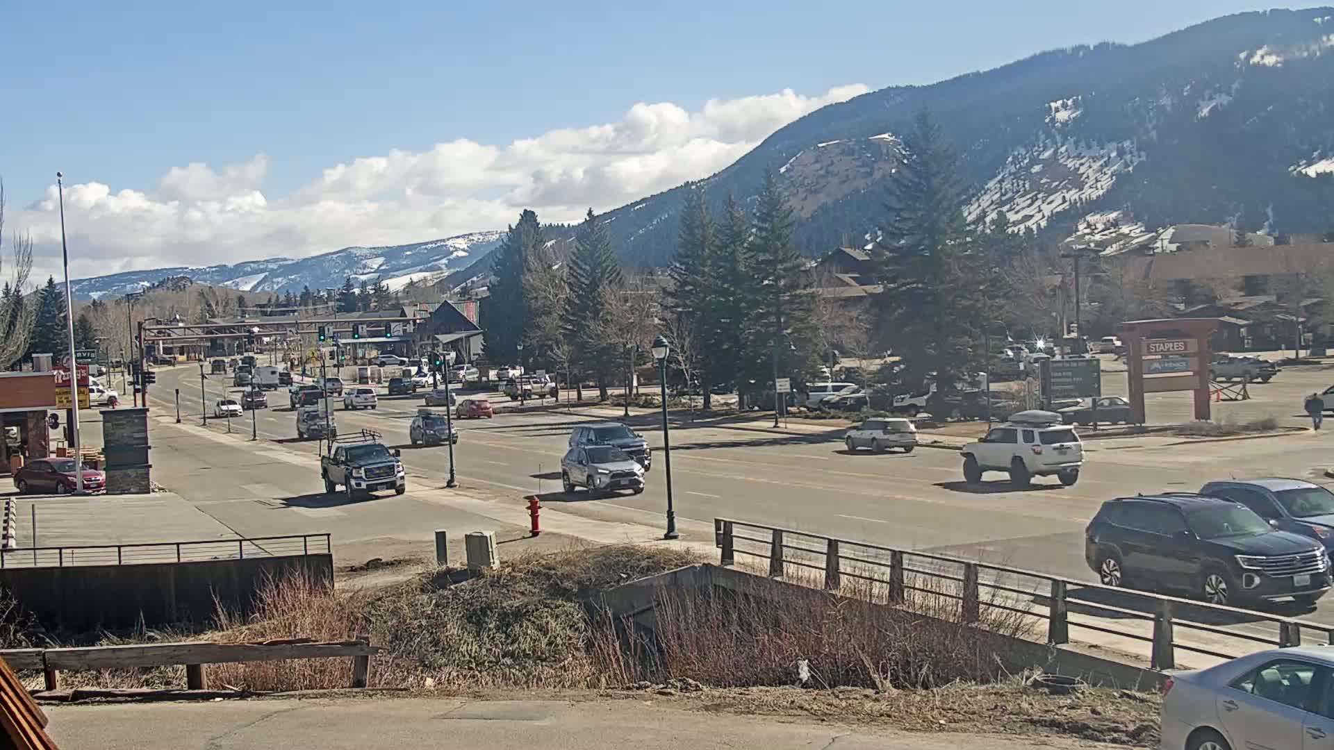 A sunny day reveals a town's main street with cars driving past buildings and snow-capped mountains in the background.
