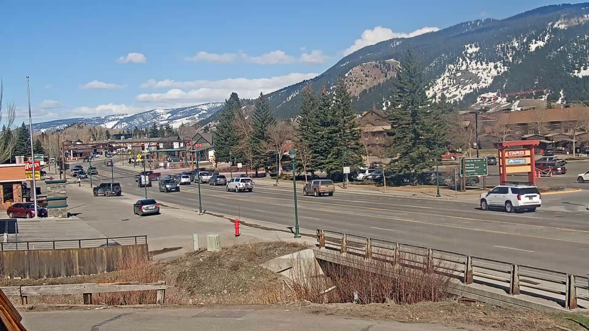 A sunny day reveals a mountain town scene with a multi-lane road, several cars, and businesses, all set against a backdrop of snow-capped mountains.