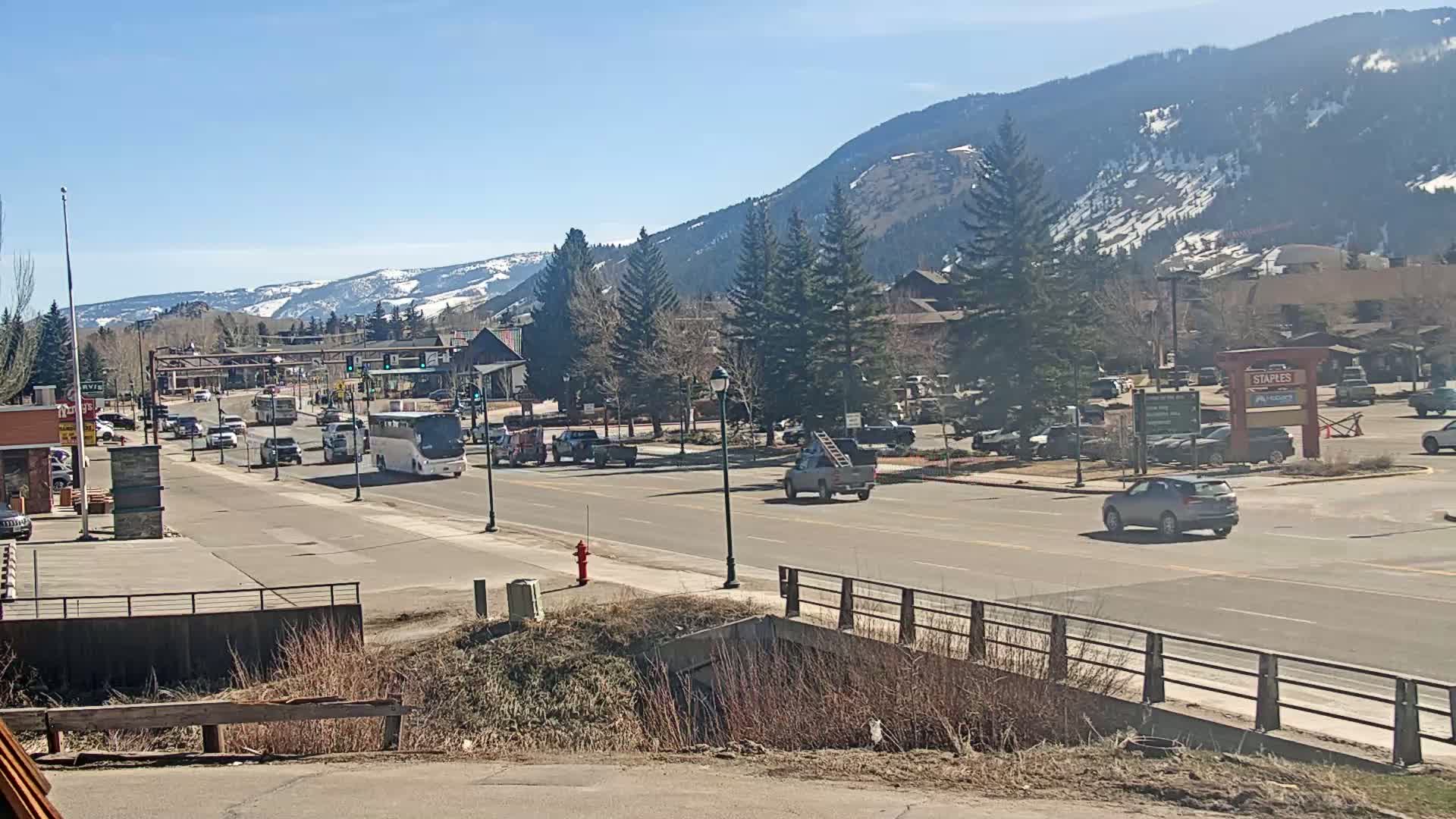 A sunny day shows a mountain town street with cars and a bus driving on a road, with snow-capped mountains in the background.