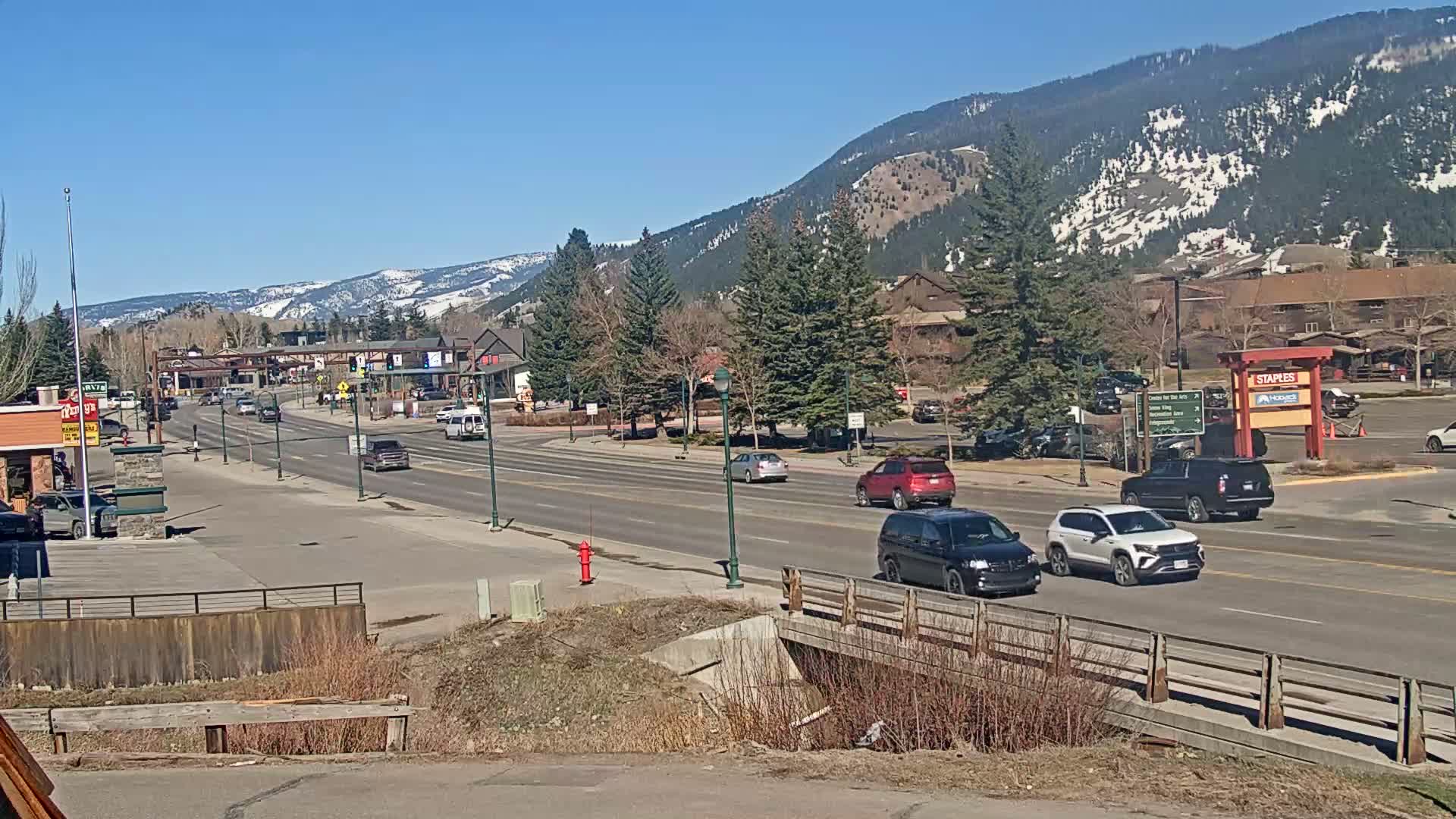 A sunny day reveals a paved road lined with buildings and trees, leading towards snow-capped mountains in the background.
