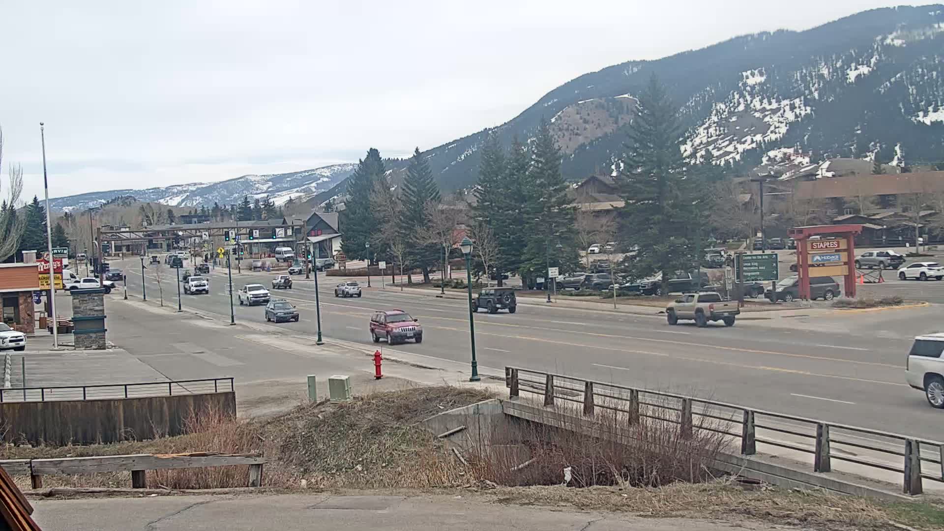 A partly cloudy day reveals a mountain town street scene with moderate traffic, featuring a two-lane road, buildings, and snow-capped mountains in the background.