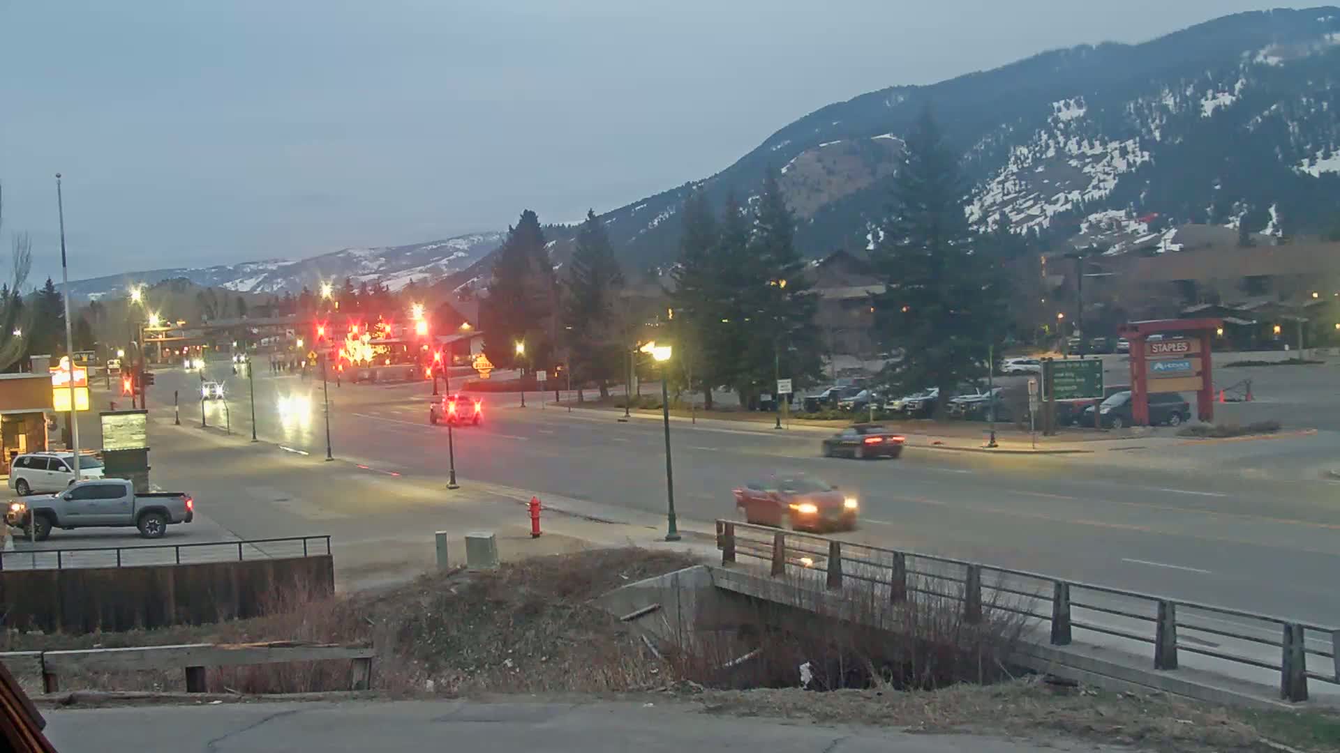 A twilight view of a multi-lane road in a town nestled against snow-dusted mountains, with several vehicles traveling in both directions.