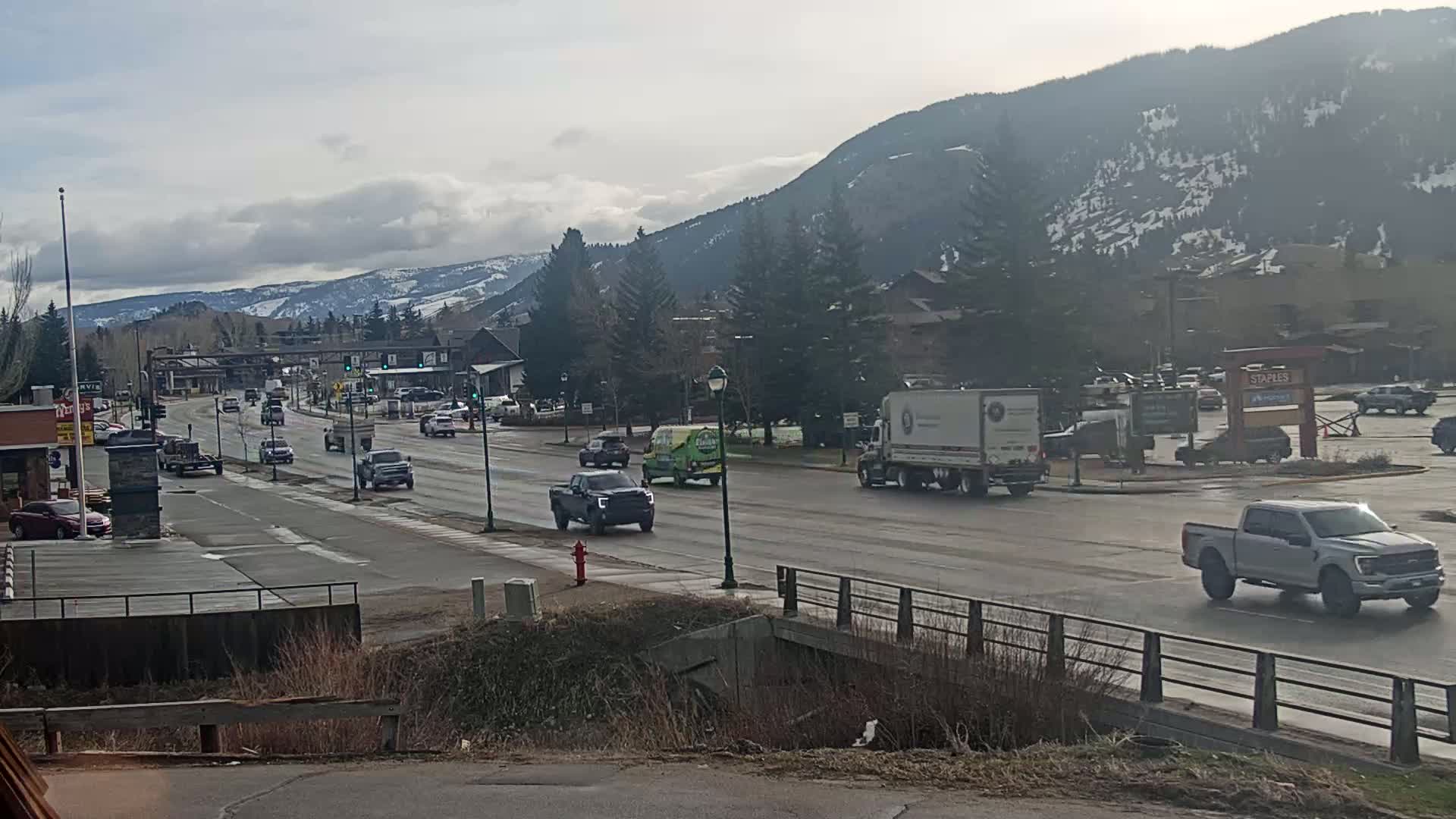 A wet, multi-lane road with several vehicles, including trucks and cars, stretches towards snow-capped mountains under a partly cloudy sky.