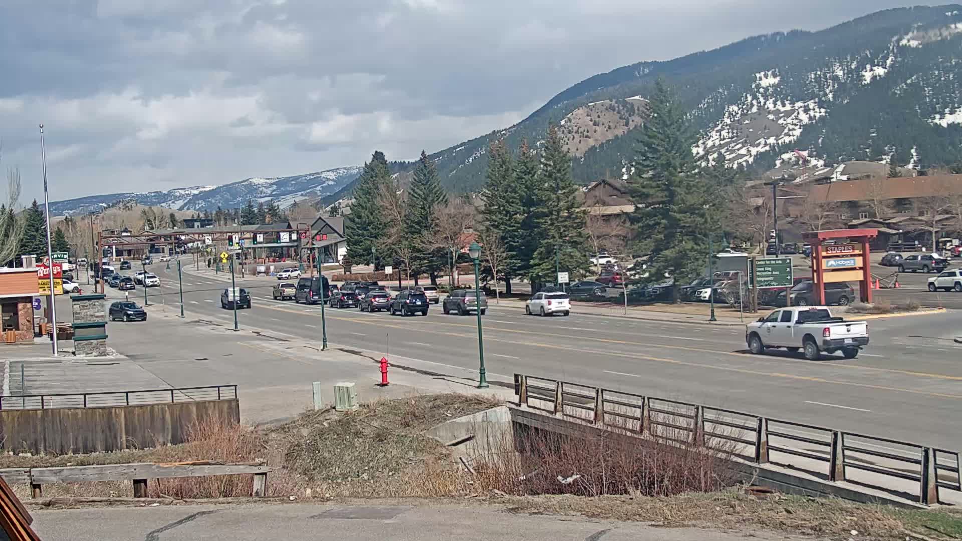 A partly cloudy day reveals a mountain town street scene with moderate traffic, lined with buildings and trees, and mountains in the background.