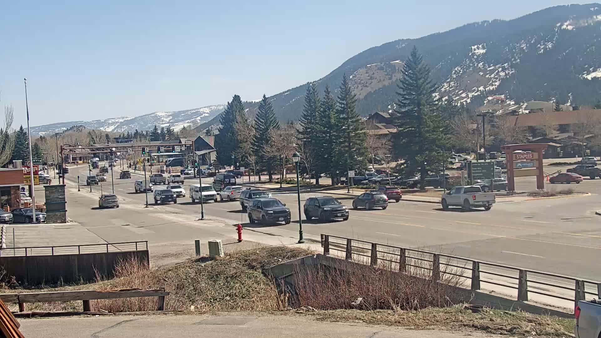 A sunny day reveals a mountain vista behind a street scene of cars driving through a town with evergreen trees and buildings.