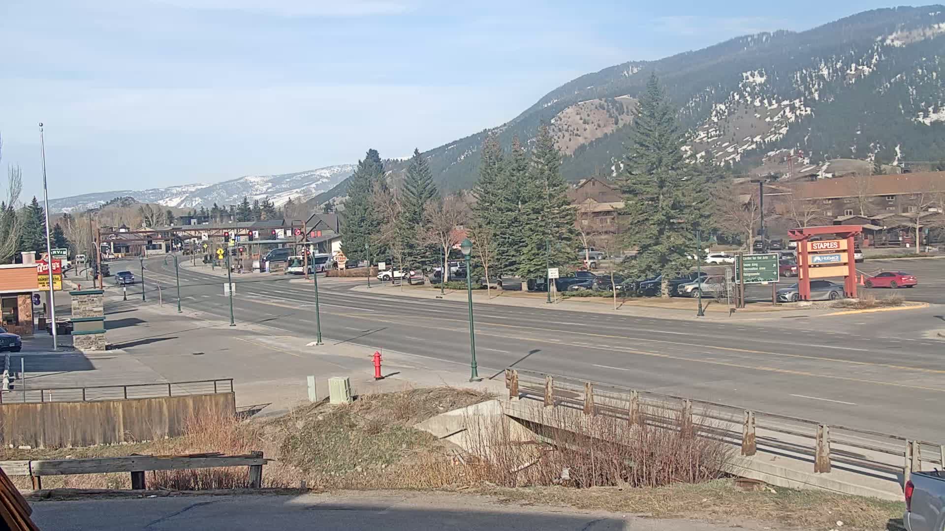 A sunny day reveals a long, relatively empty road in a town nestled at the base of snow-capped mountains, with sparse traffic and buildings visible on both sides of the road.