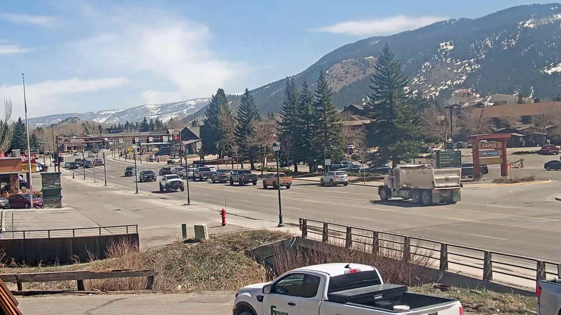 A sunny day shows a town street with moderate traffic, lined with evergreen trees and buildings, nestled against a snow-capped mountain range in the background.