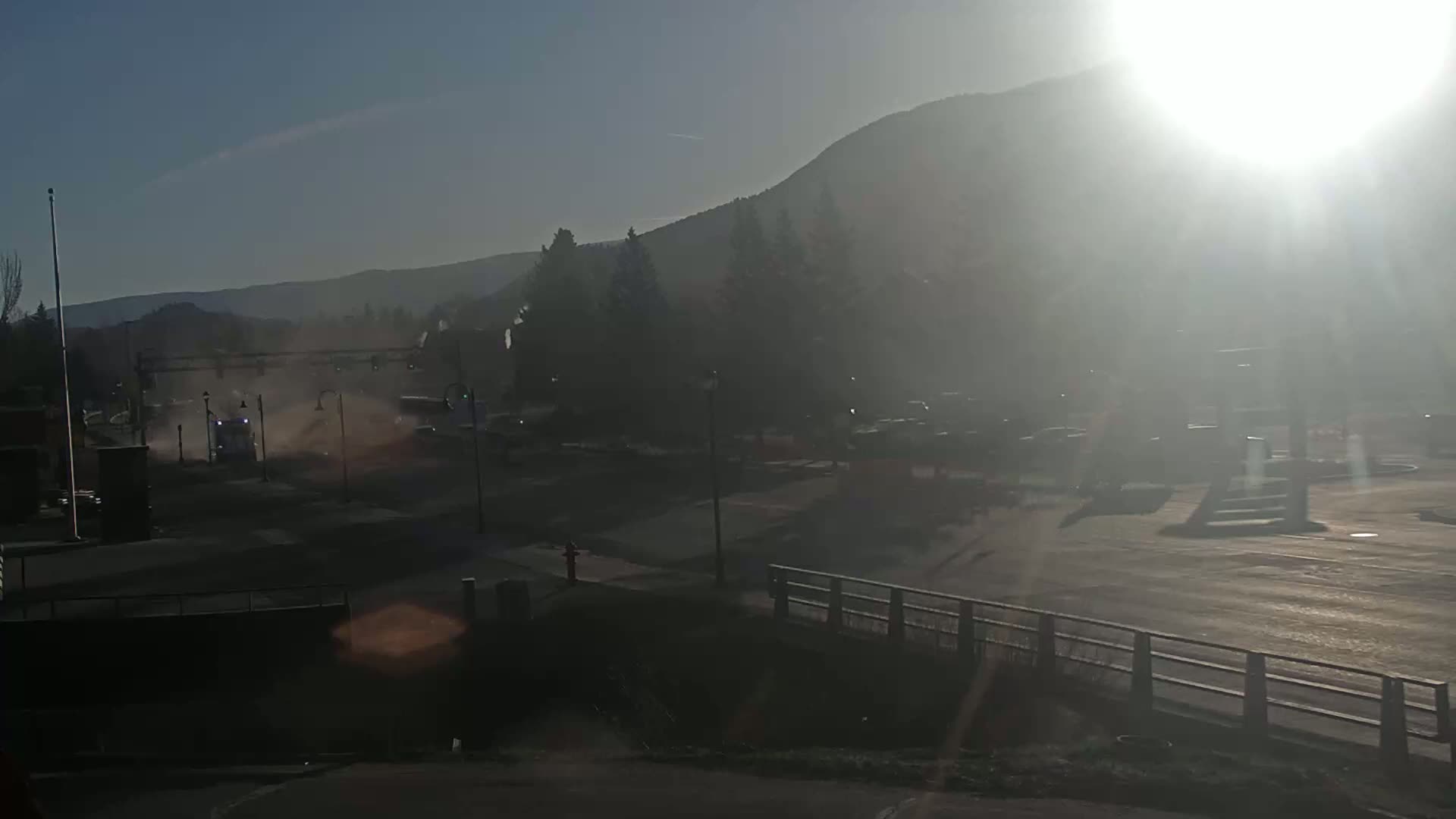 A hazy, sunny day reveals a street scene with a vehicle creating a dust cloud near a mountain backdrop.