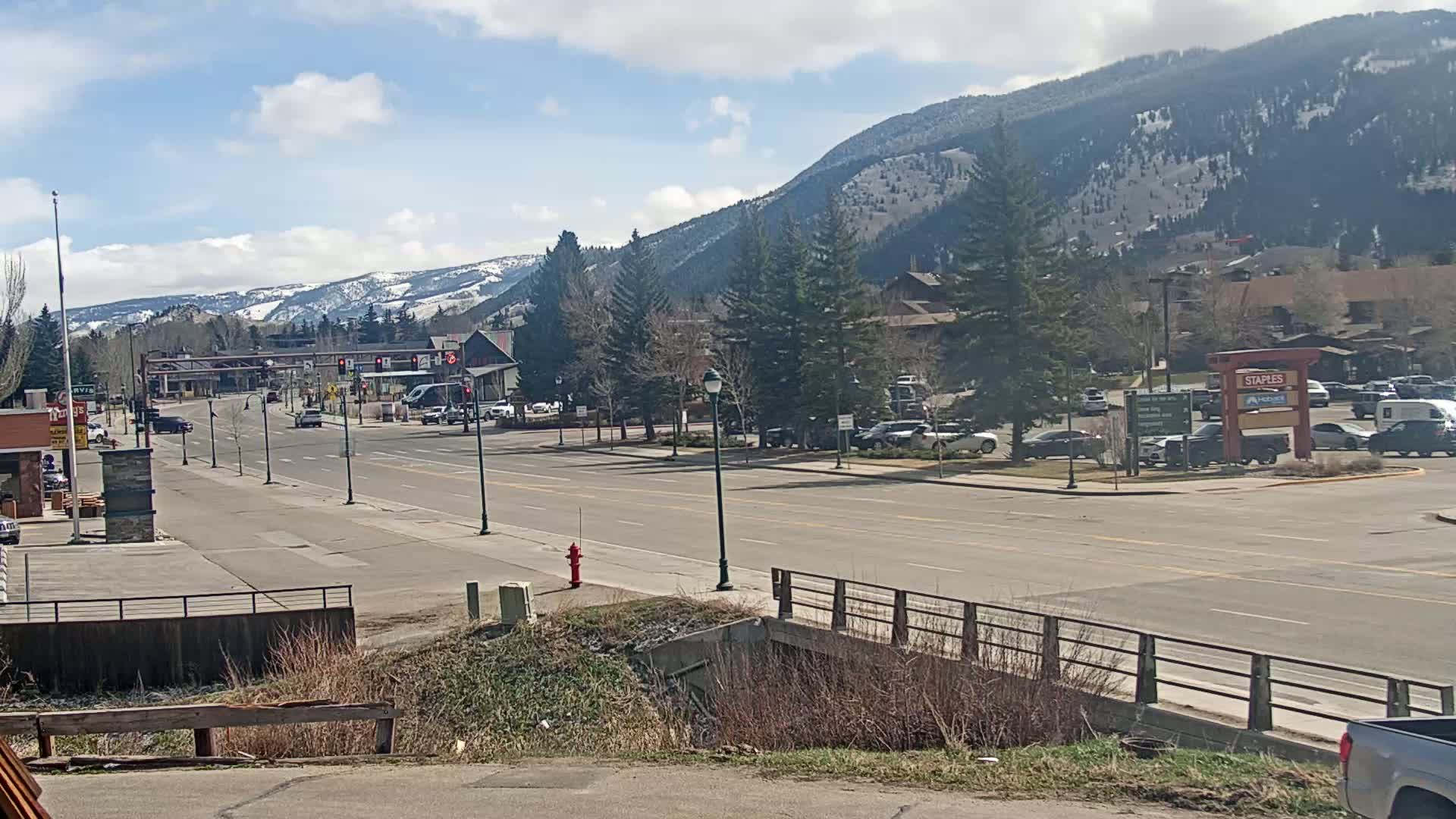 A mostly empty street in a mountain town on a partly sunny day, with snow-capped mountains in the background and cars parked along the sides of the street.
