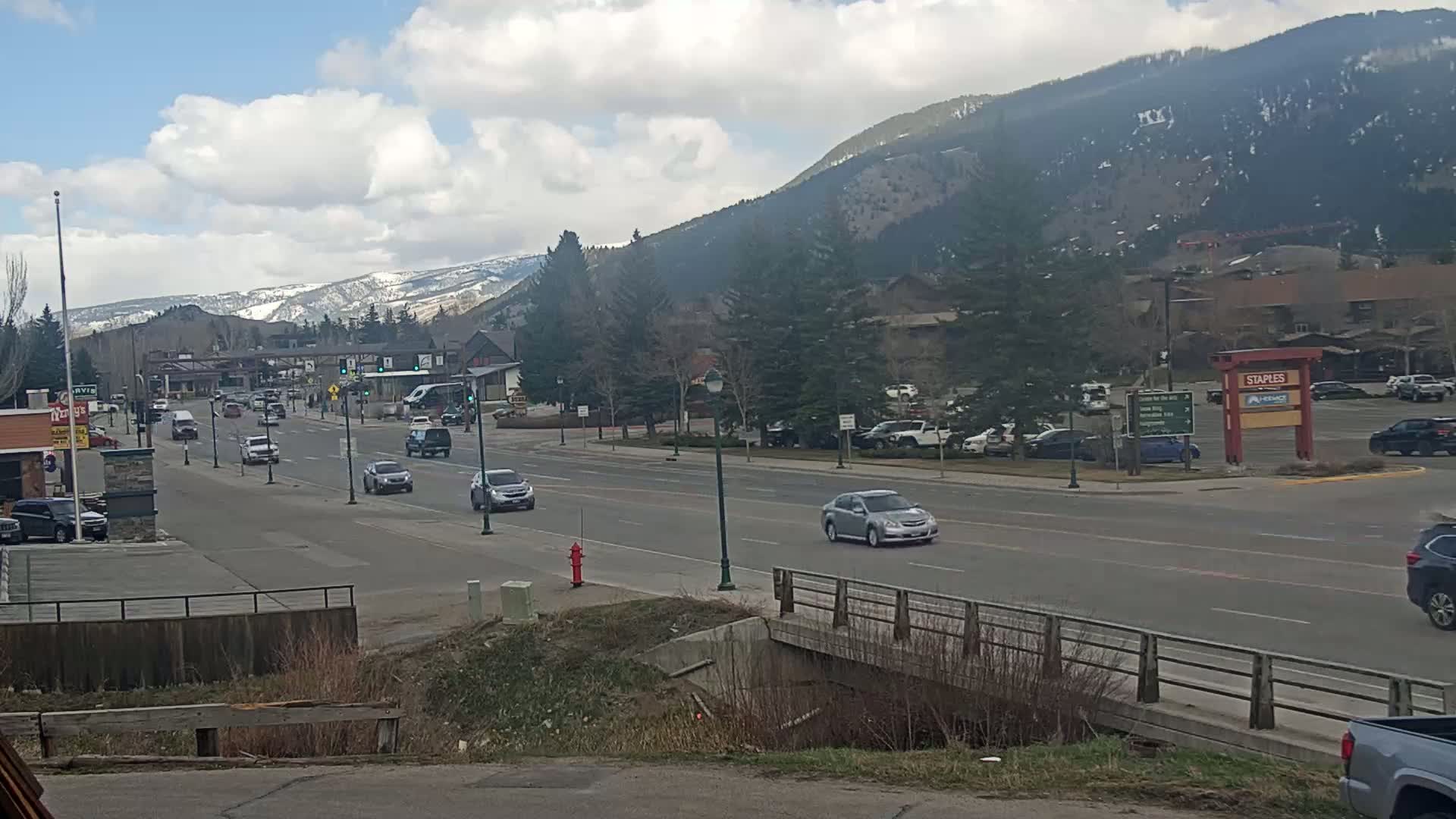 A partly cloudy day reveals a mountain range in the background, with a roadway flanked by buildings and trees in the foreground, featuring several cars driving on a street.
