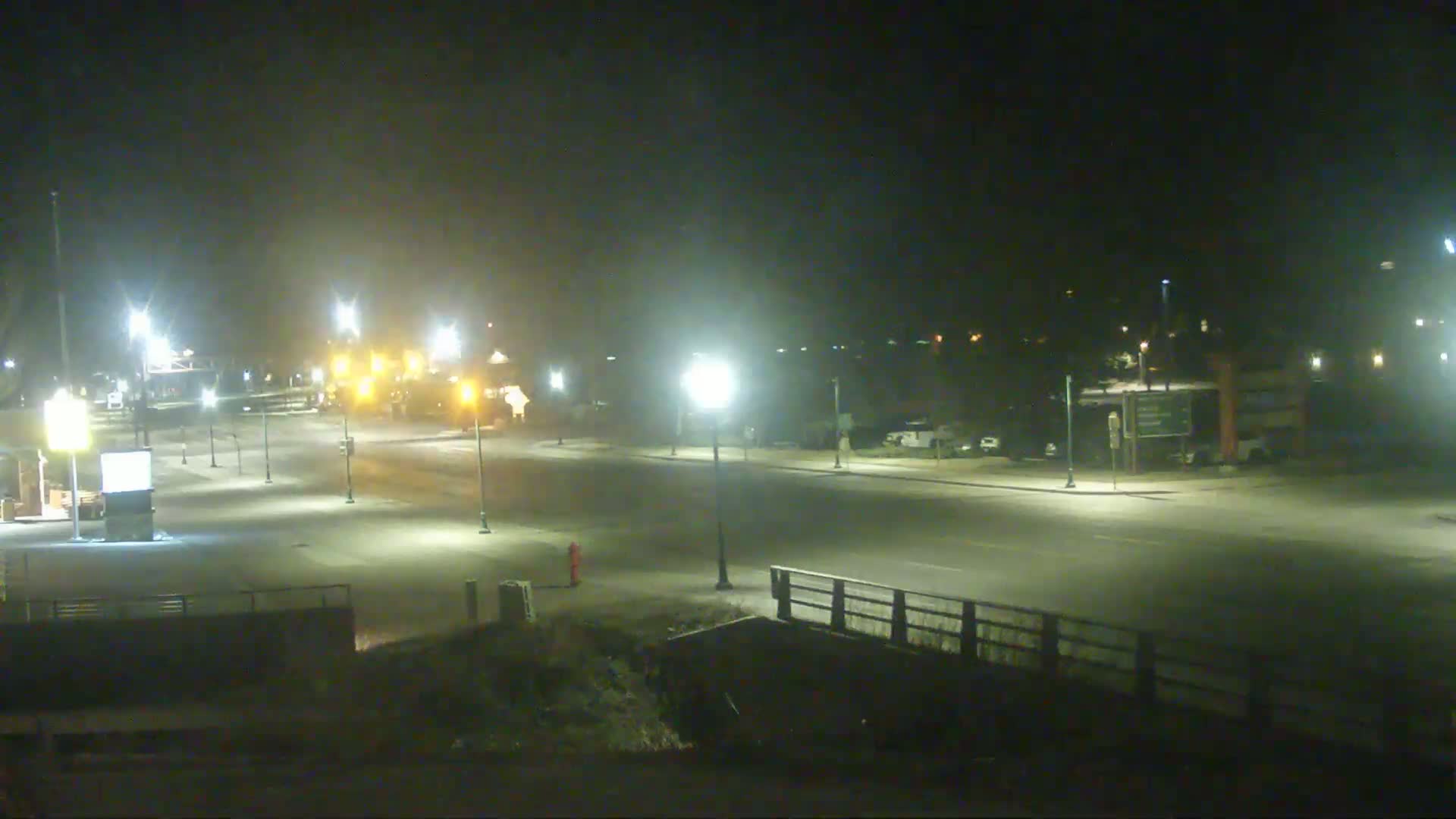 A mostly empty, well-lit roadway at night, under hazy conditions, with a few parked cars visible in the distance.