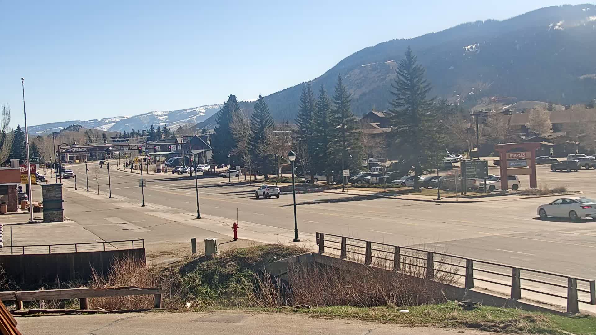 A mostly sunny day reveals a long road leading to snow-capped mountains, lined with buildings, parking lots, and sparsely populated with vehicles.