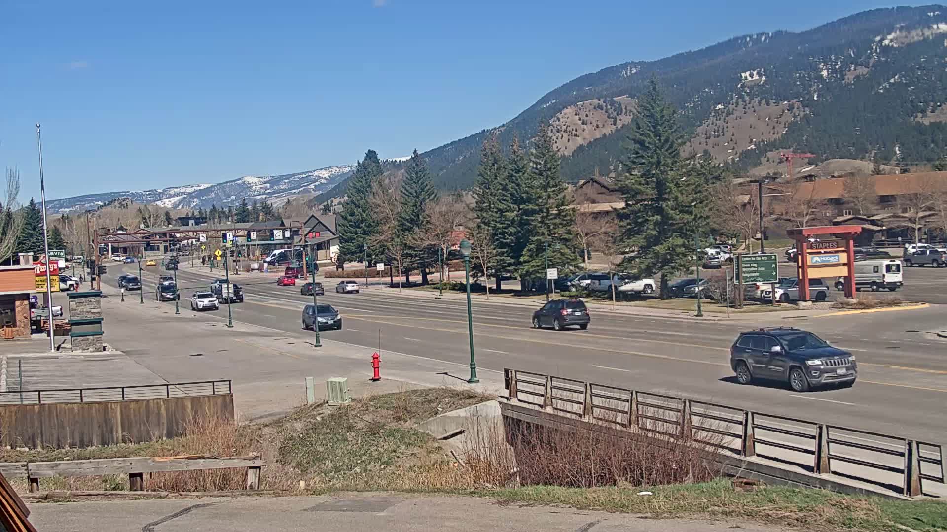 A sunny day reveals a town's main street, lined with buildings, trees, and parked cars, leading towards snow-capped mountains in the distance.