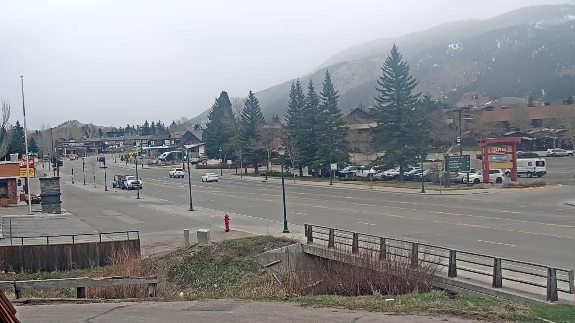 A hazy, overcast day reveals a multi-lane road with sparse traffic passing through a town nestled at the foot of a mountain range, lined with buildings and evergreens.