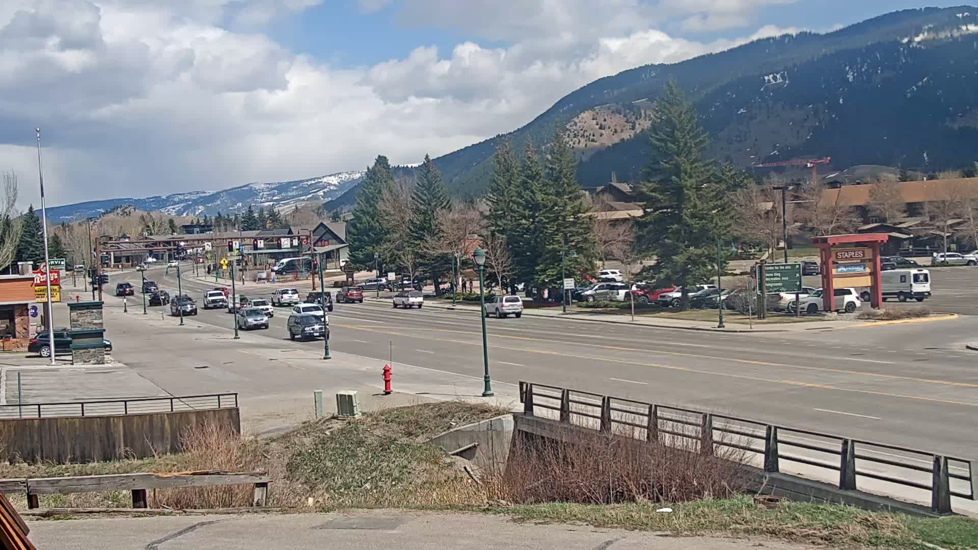 A partly cloudy day reveals a mountain view overlooking a town's main street with cars driving on a multi-lane road, parked cars lining the sides, and various buildings and trees in the background.