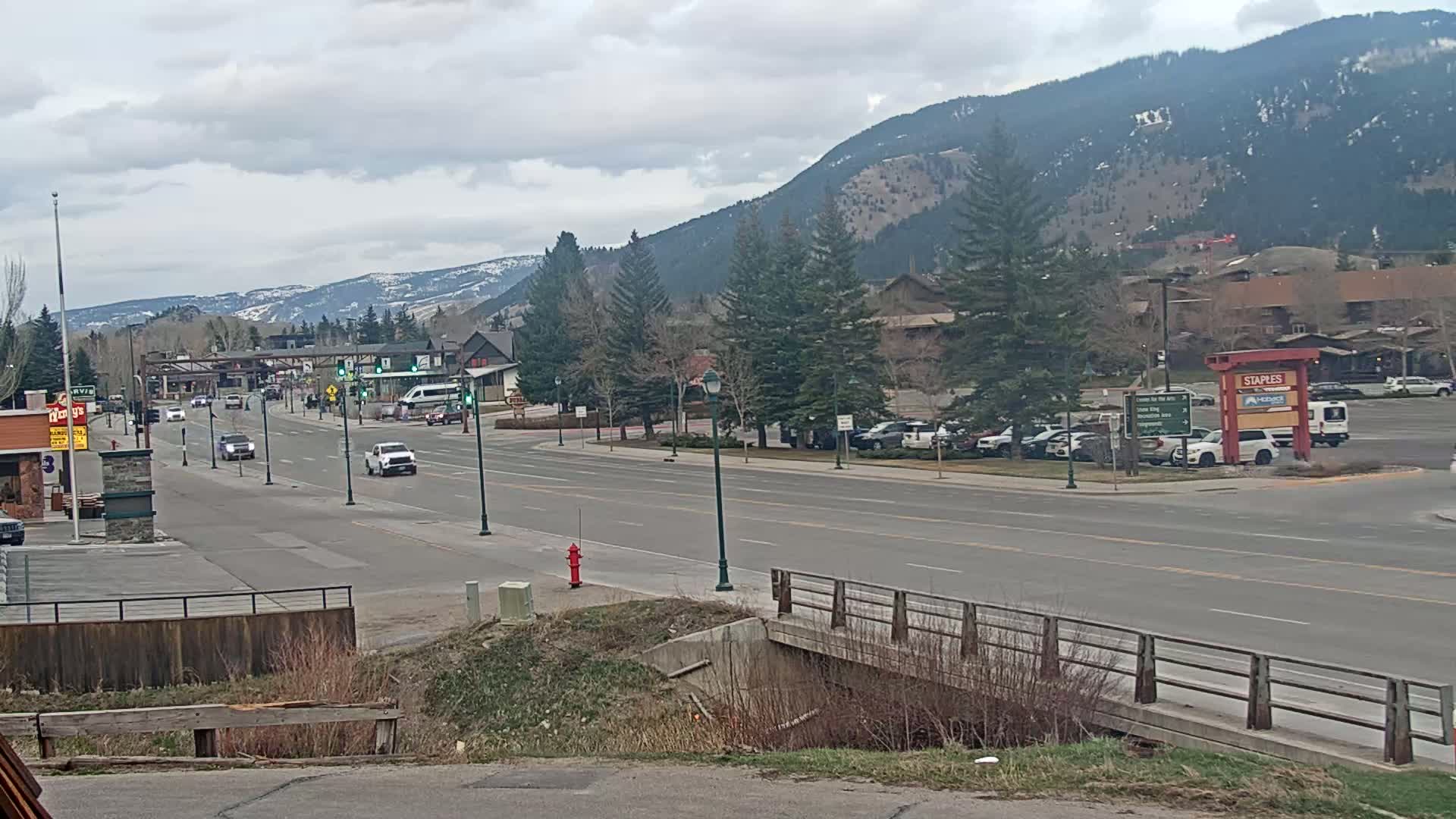A multi-lane road winds through a town nestled against a mountain range under a cloudy sky, with sparse traffic and several buildings visible.