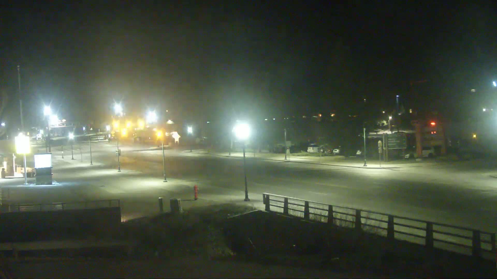 A mostly empty, snow-covered street is lit by streetlights at night, with a few vehicles visible in the distance under hazy conditions.