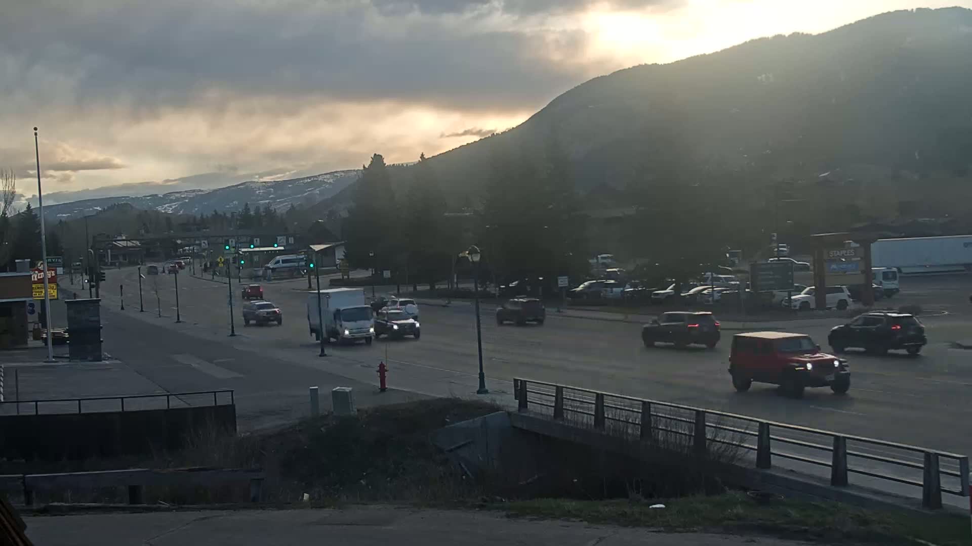 Cars drive on a road that passes through a town nestled at the base of a mountain range under a partly cloudy sunset sky.