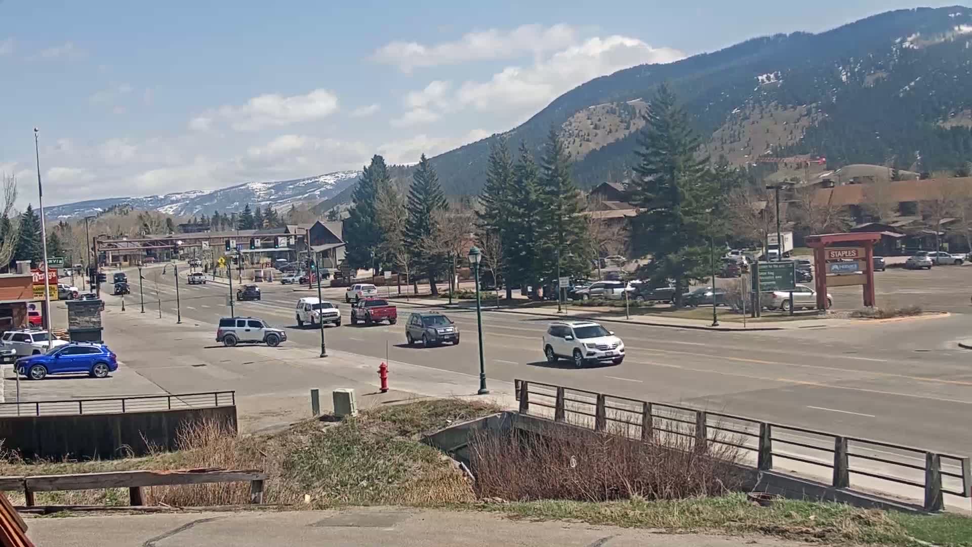 A sunny day reveals a mountain town street scene with several vehicles driving on a road that passes in front of buildings and parking lots, with snow-capped mountains visible in the distance.