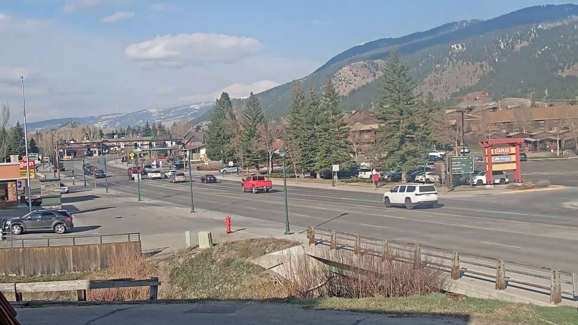 A sunny day reveals a multi-lane road with sparse traffic passing through a town nestled at the base of snow-capped mountains.
