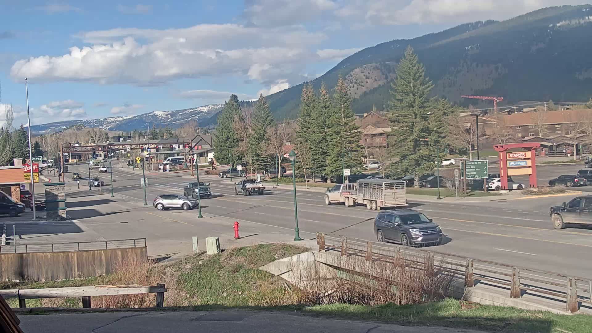 A partly cloudy day reveals a mountain town street scene with several cars and trucks driving on a multi-lane road, bordered by buildings and trees, under a bright blue sky.