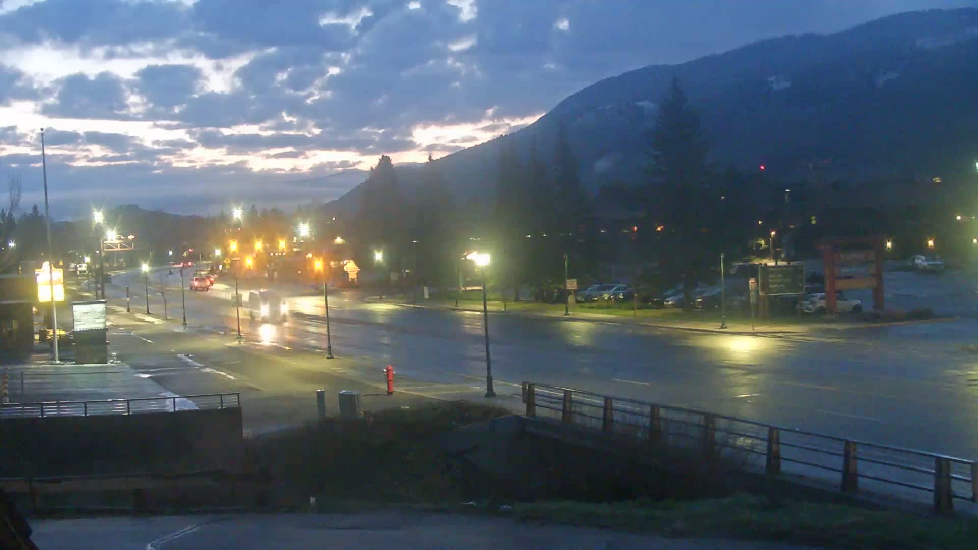 A wet, dimly lit street scene at dawn shows a few cars driving past buildings and parking lots beneath a cloudy sky and mountains in the background.