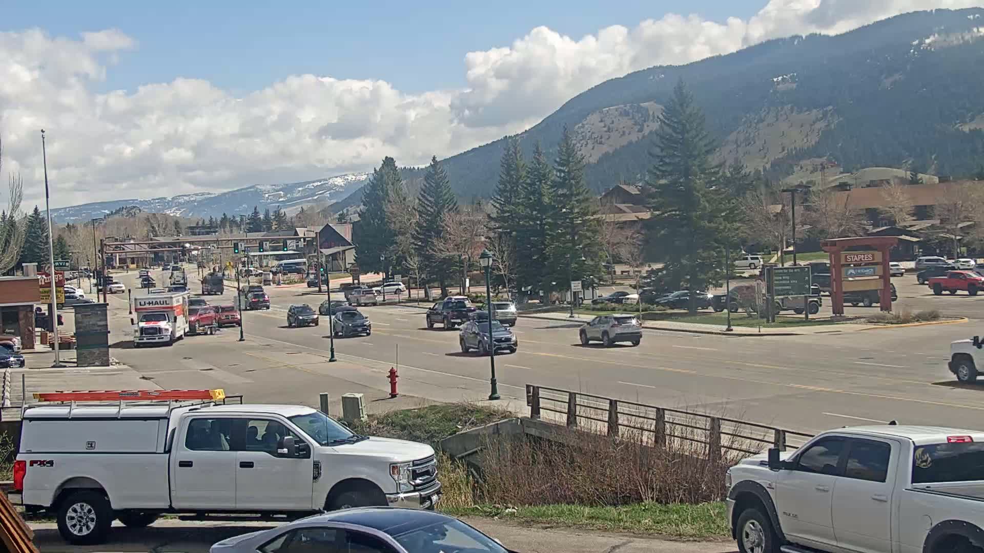 A sunny day with partly cloudy skies shows a mountain town's main street with several vehicles, including trucks and cars, driving in both directions.