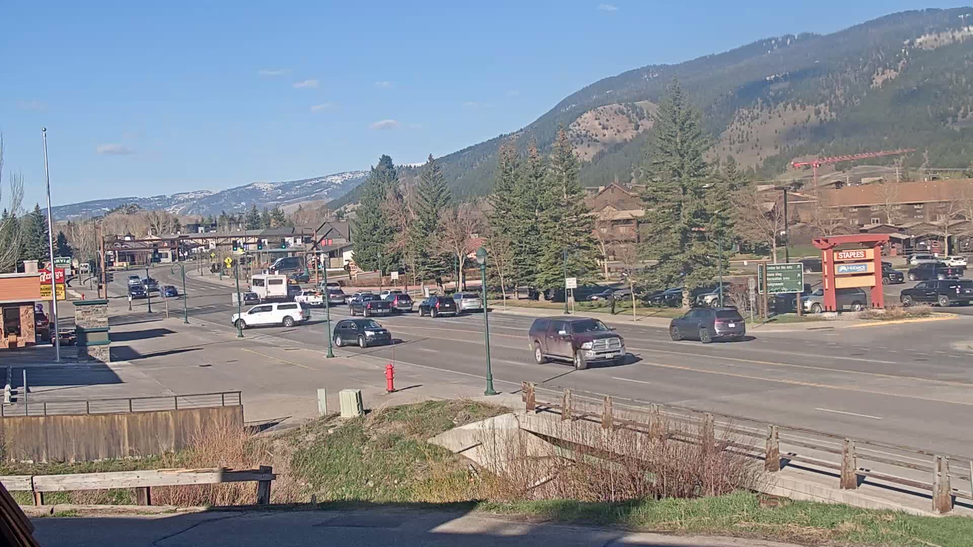A sunny day reveals a two-lane road lined with cars, passing through a town nestled against a mountain range under a clear blue sky.