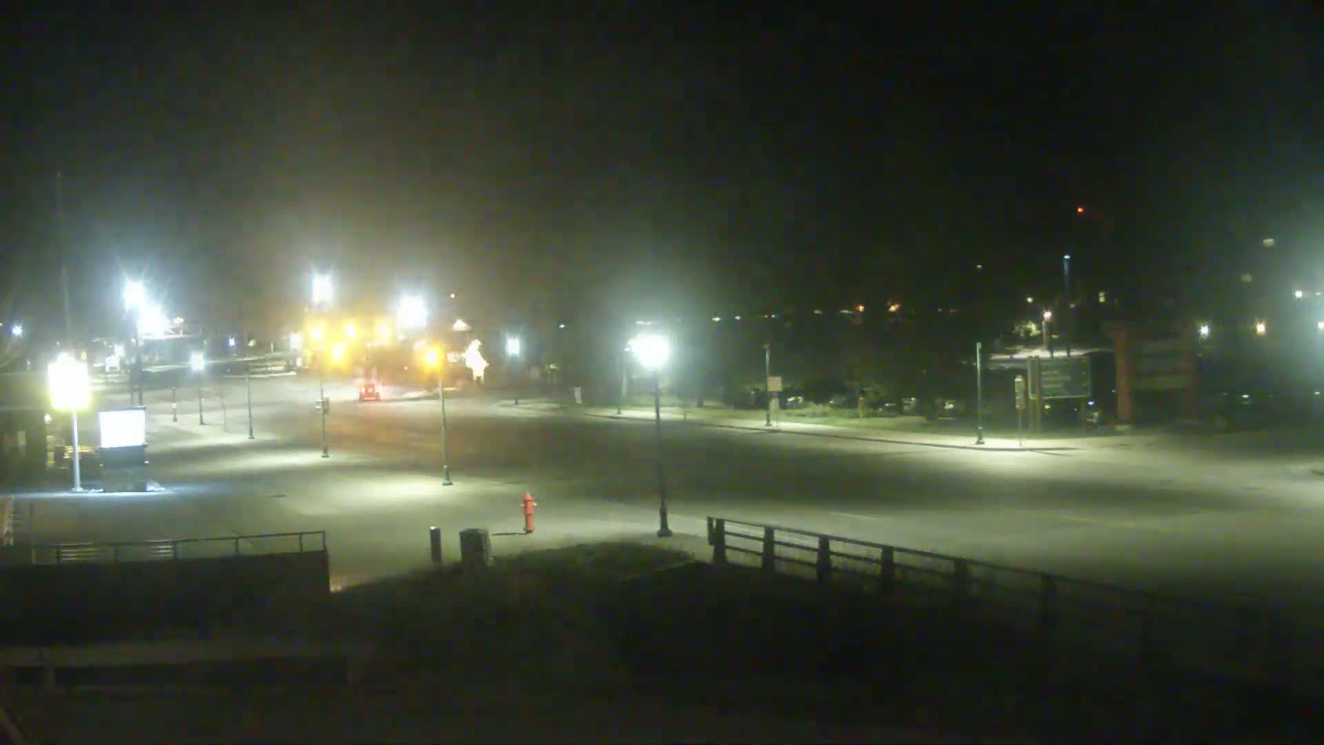 A nighttime view of a mostly empty road with streetlights, a few vehicles, and some vegetation visible in the background under hazy conditions.