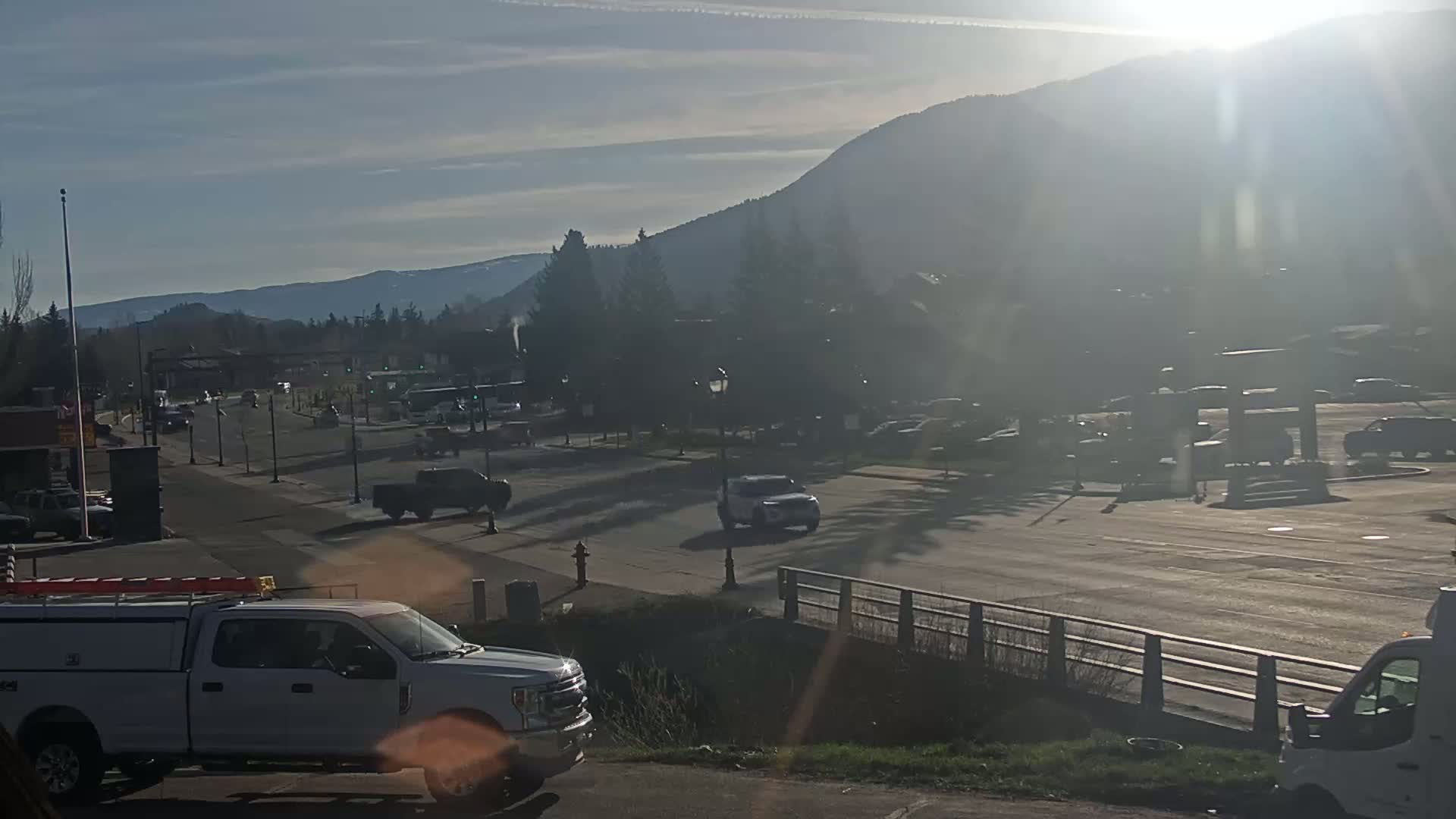 A sunny day reveals a mountain view behind a town's street scene, where several vehicles are parked and driving along a road with a low fence.