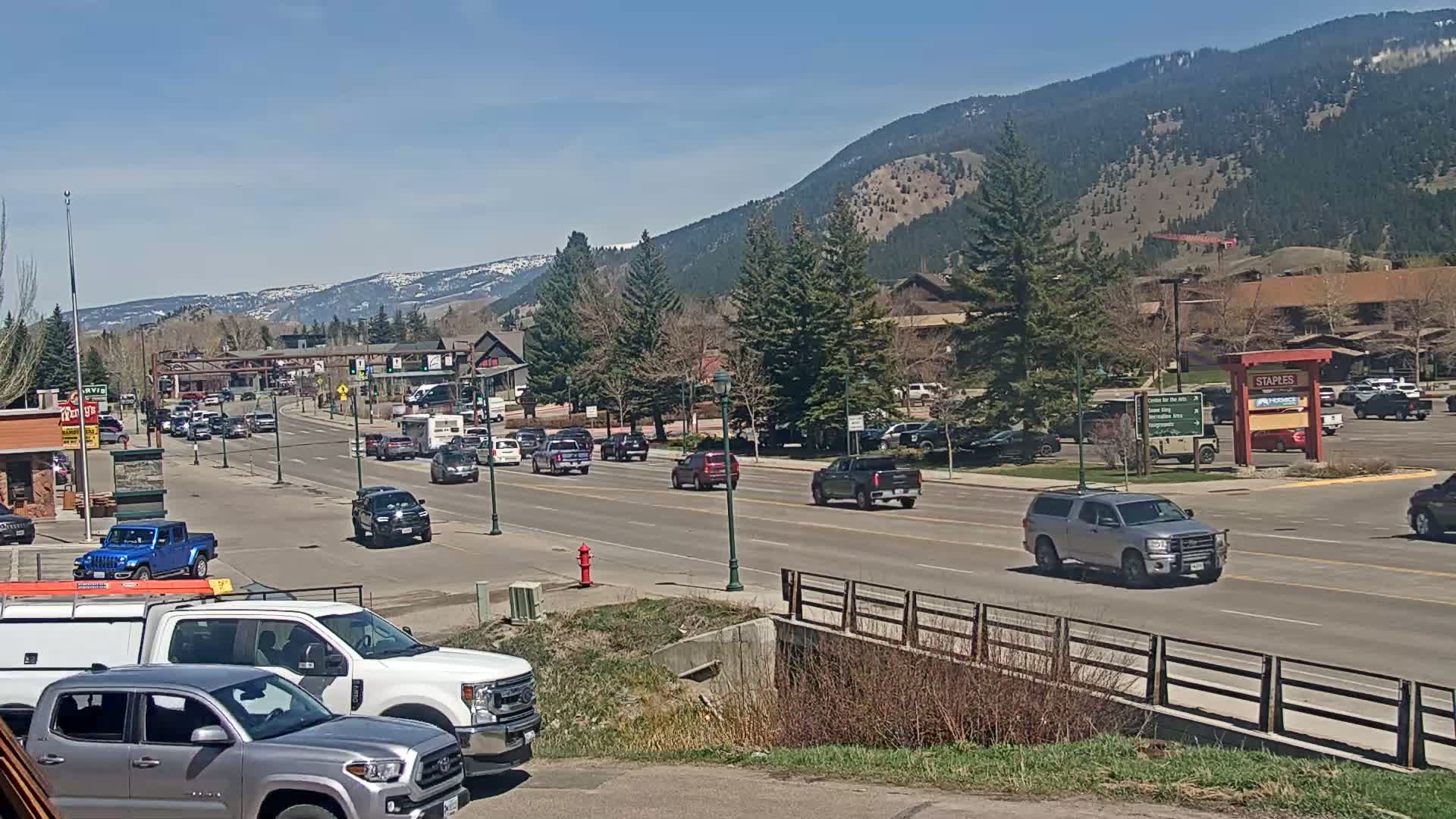 A sunny day reveals a busy street scene in a town nestled against a mountain range, with cars driving on a road lined with trees and buildings.