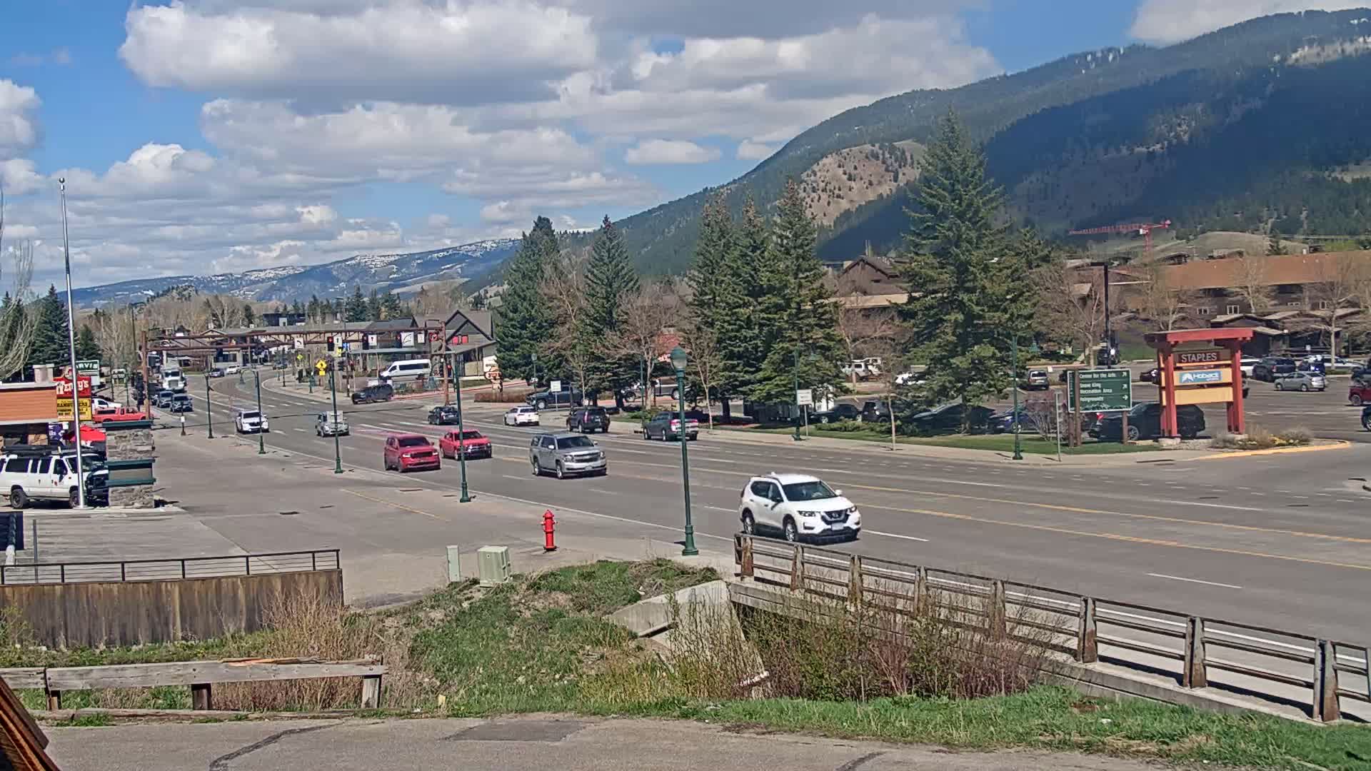 A partly cloudy day reveals a mountain town street scene with several cars driving along a road lined with trees and buildings, a bridge in the foreground, and snow-capped mountains in the distance.