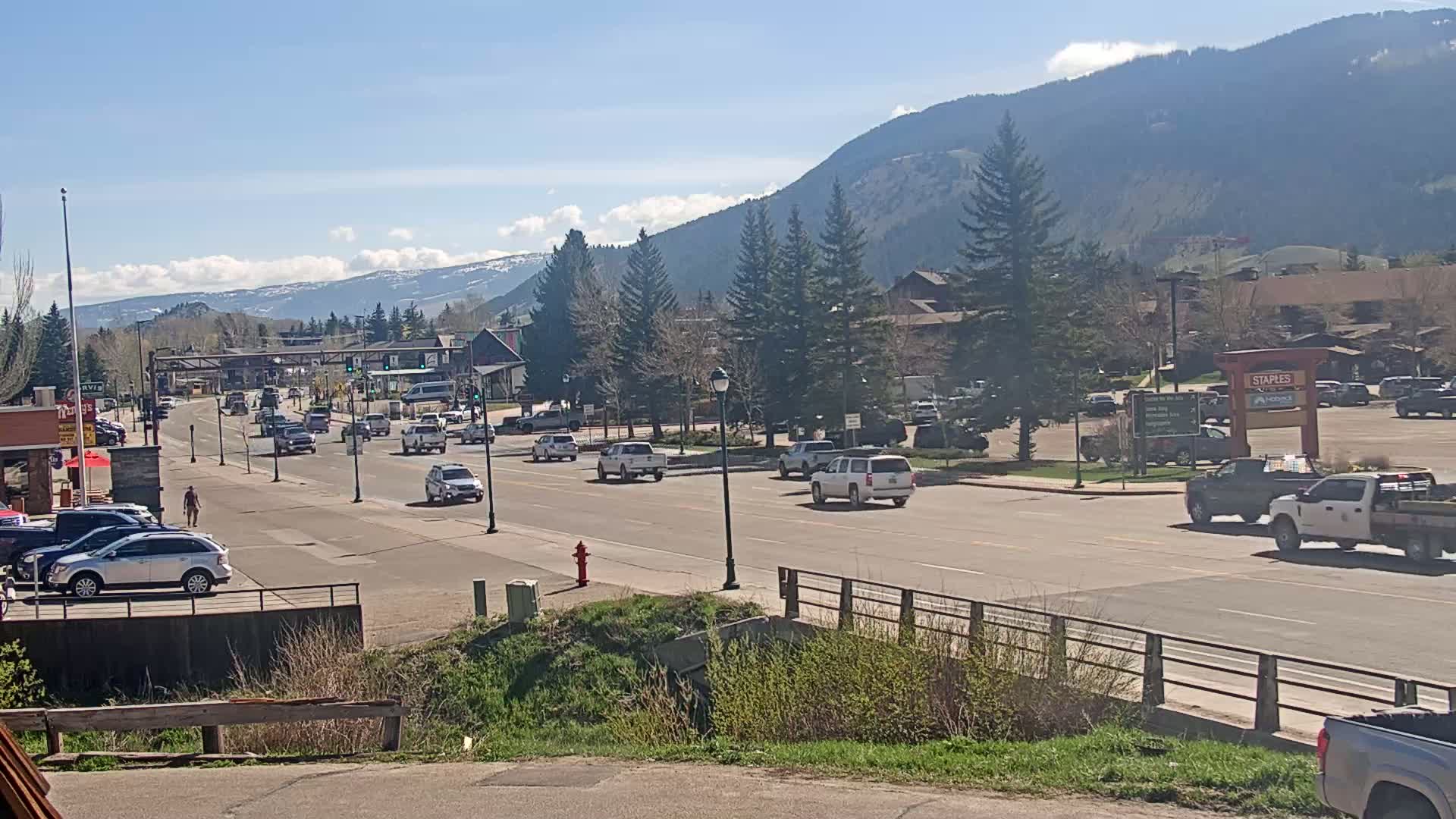 A sunny day reveals a mountain town's main street with moderate traffic, lined with buildings and trees, mountains in the background.