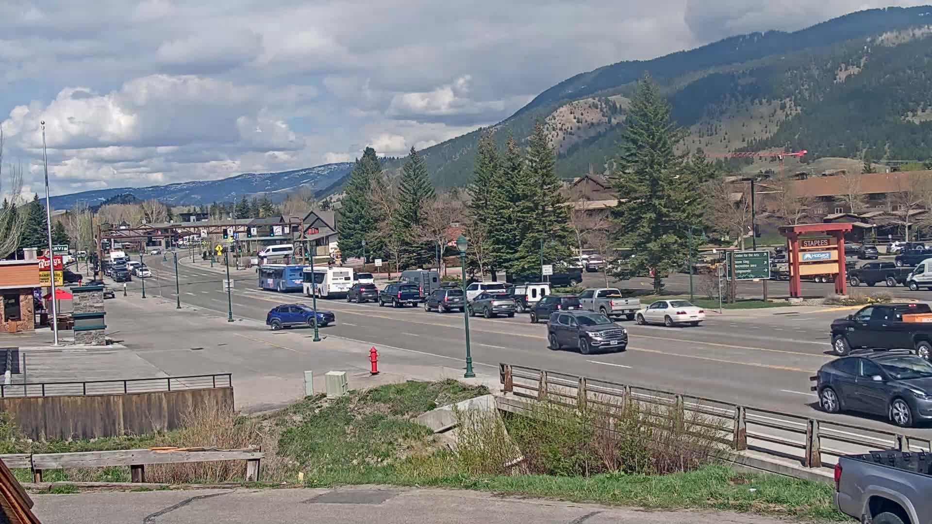 A partly cloudy day reveals a mountain town's main street, with moderate traffic including cars and buses, and a small bridge in the foreground.