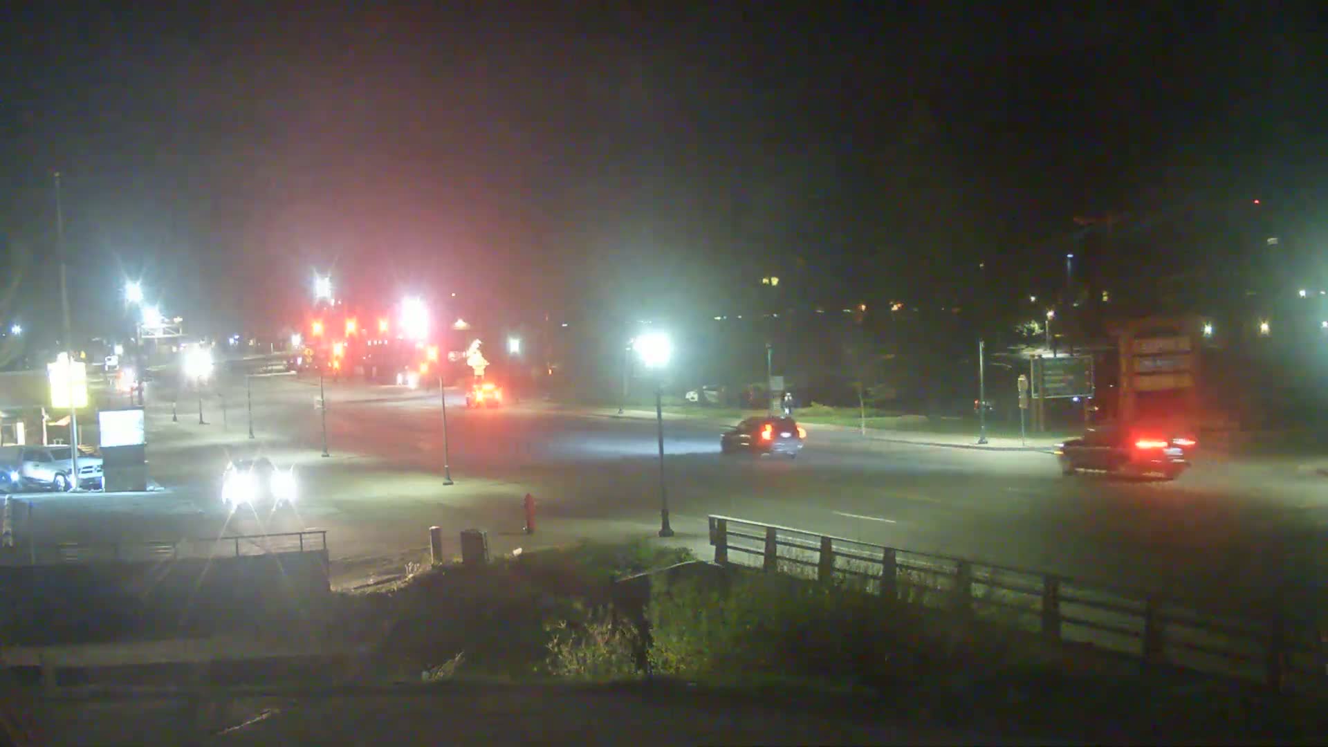 A nighttime scene shows a few vehicles traveling on a road illuminated by streetlights, with a hazy or foggy atmosphere.