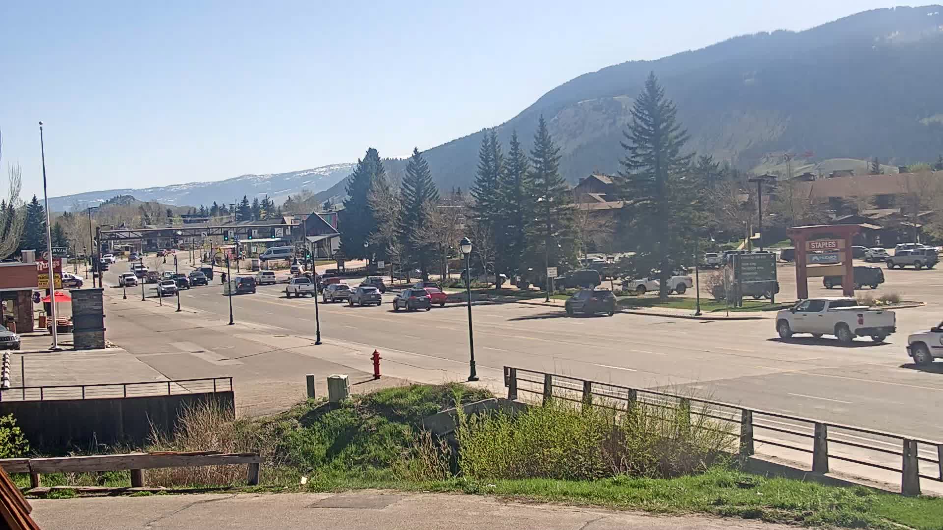 A sunny day reveals a multi-lane road lined with cars, passing through a town nestled at the foot of a mountain range.