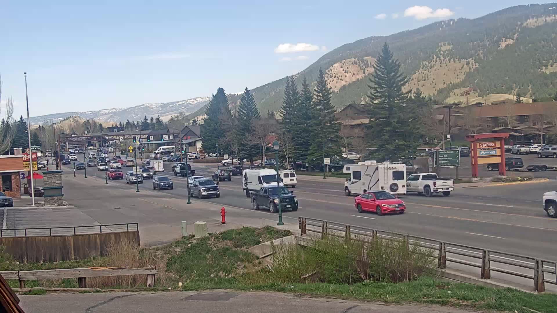 A sunny day reveals a two-lane road with moderate traffic flowing past shops and buildings nestled at the base of a mountain range covered in evergreens.