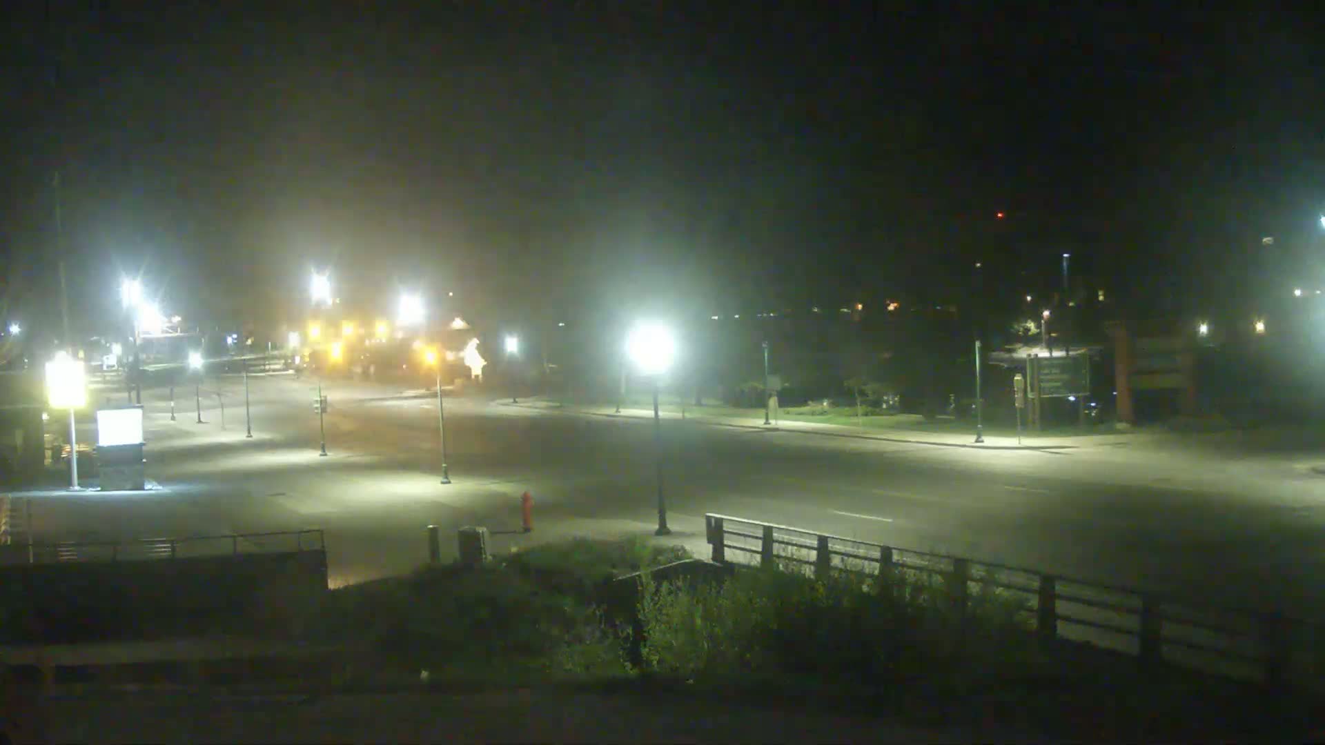 A mostly empty, lightly fog-shrouded road at night is illuminated by streetlights.