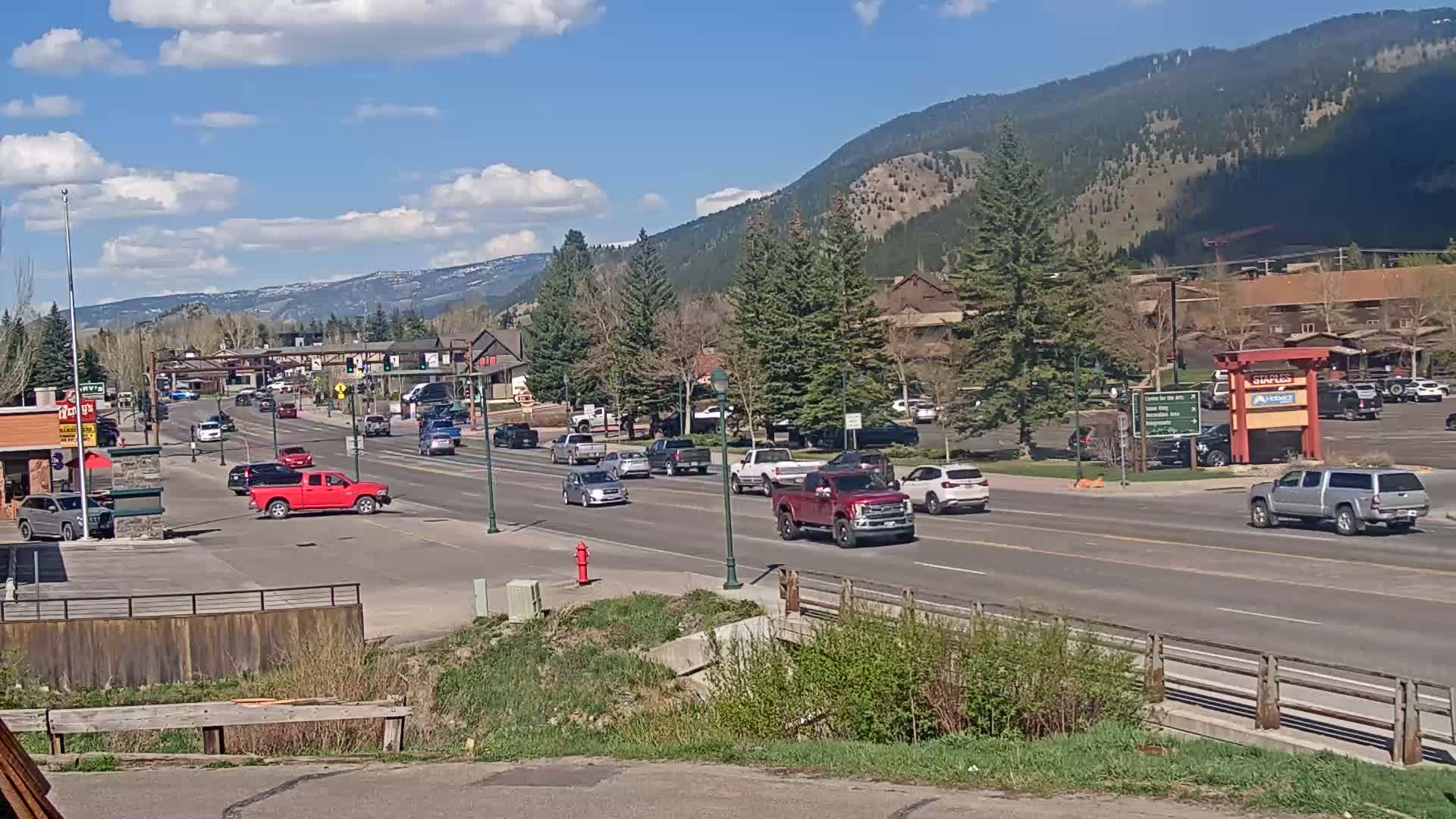 A sunny day reveals a multi-lane road in a mountain town, with cars driving past buildings and trees, and mountains visible in the background.