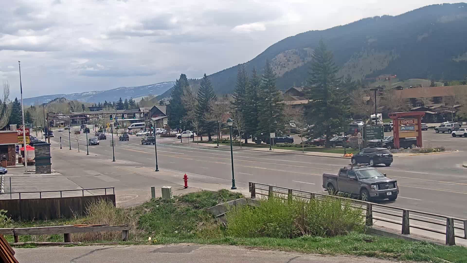 A partly cloudy day shows a multi-lane road with light traffic in a mountain town, with buildings and parking lots on either side, and mountains in the background.