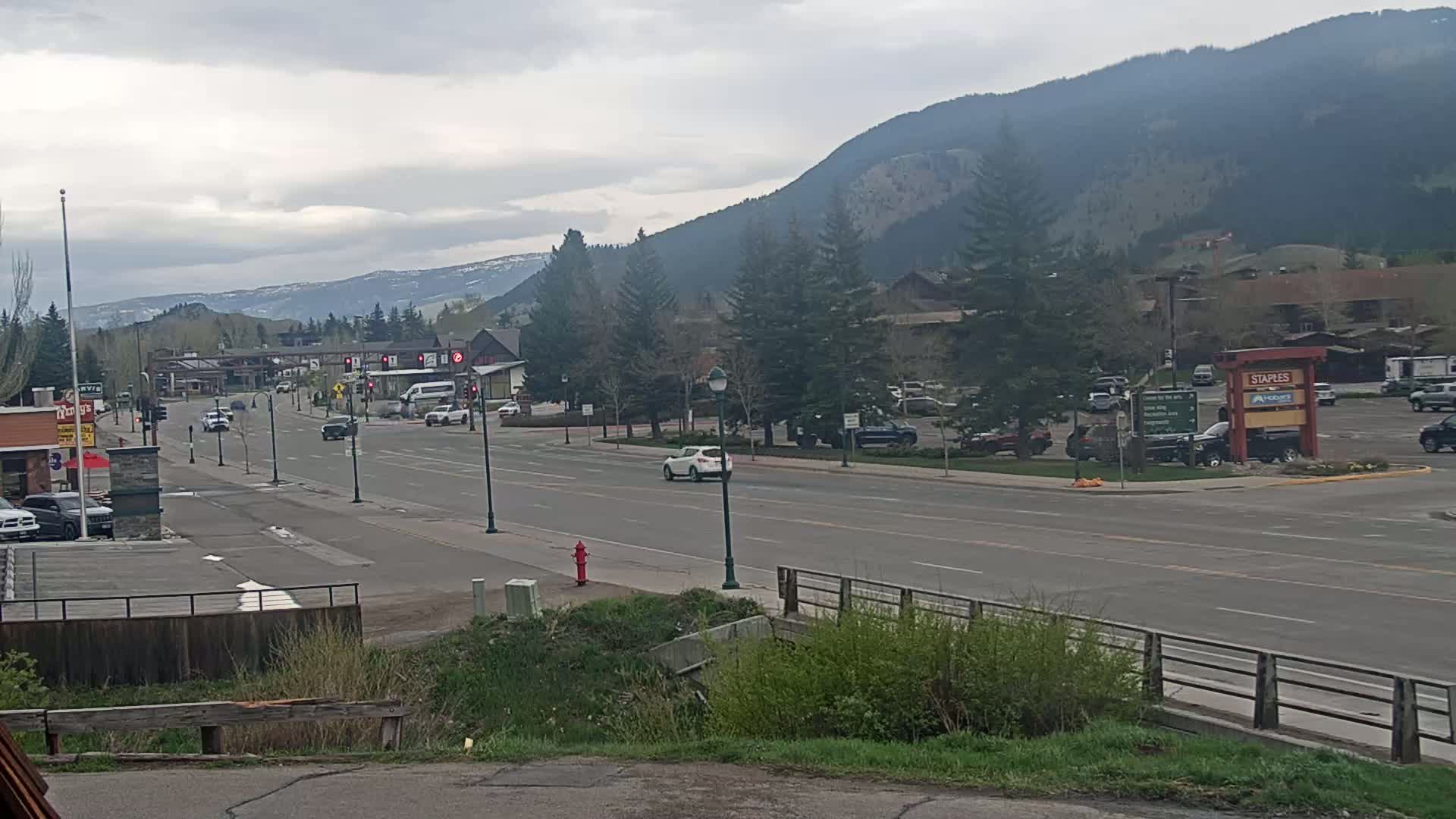 A long, relatively empty road with sparse traffic passes through a town nestled in a valley surrounded by mountains under an overcast sky.