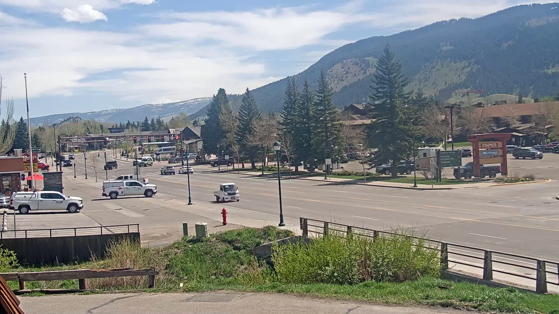 A sunny day reveals a town street scene with several vehicles, including trucks and small cars, passing through a town situated at the base of a mountain range.
