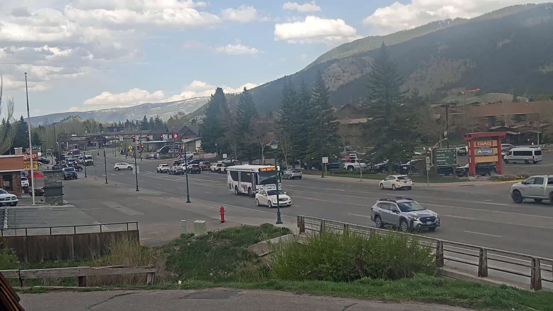 A partly cloudy day shows a town's main street with moderate traffic, including a shuttle bus, against a backdrop of mountains.