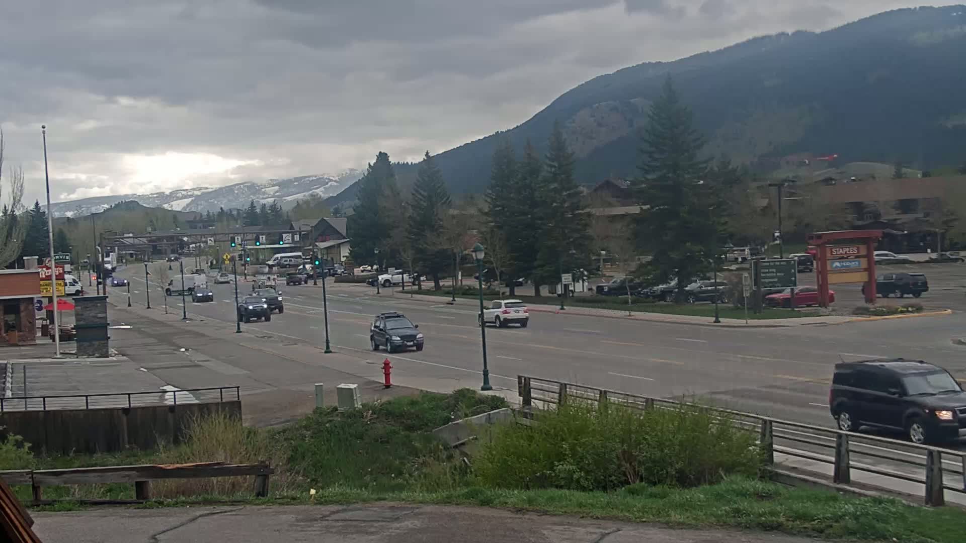 A street scene on an overcast day shows several vehicles driving along a road lined with trees and businesses, with snow-capped mountains in the background.
